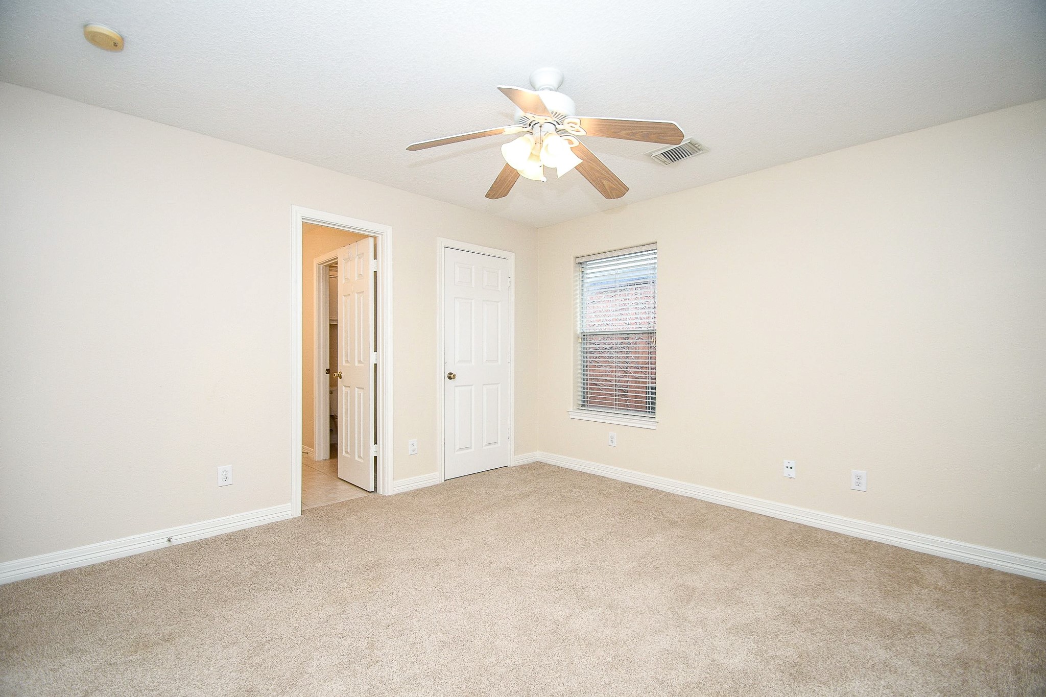This is a bright, carpeted bedroom with neutral walls, featuring a ceiling fan and a window for natural light. There is an adjacent door leading to a bathroom.