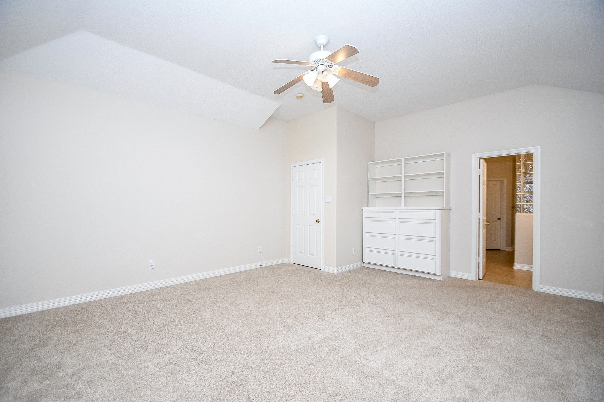 Spacious, bright room with neutral carpeting, ceiling fan, built-in shelving, and a doorway leading to an adjoining room. Ideal for a bedroom or office space.