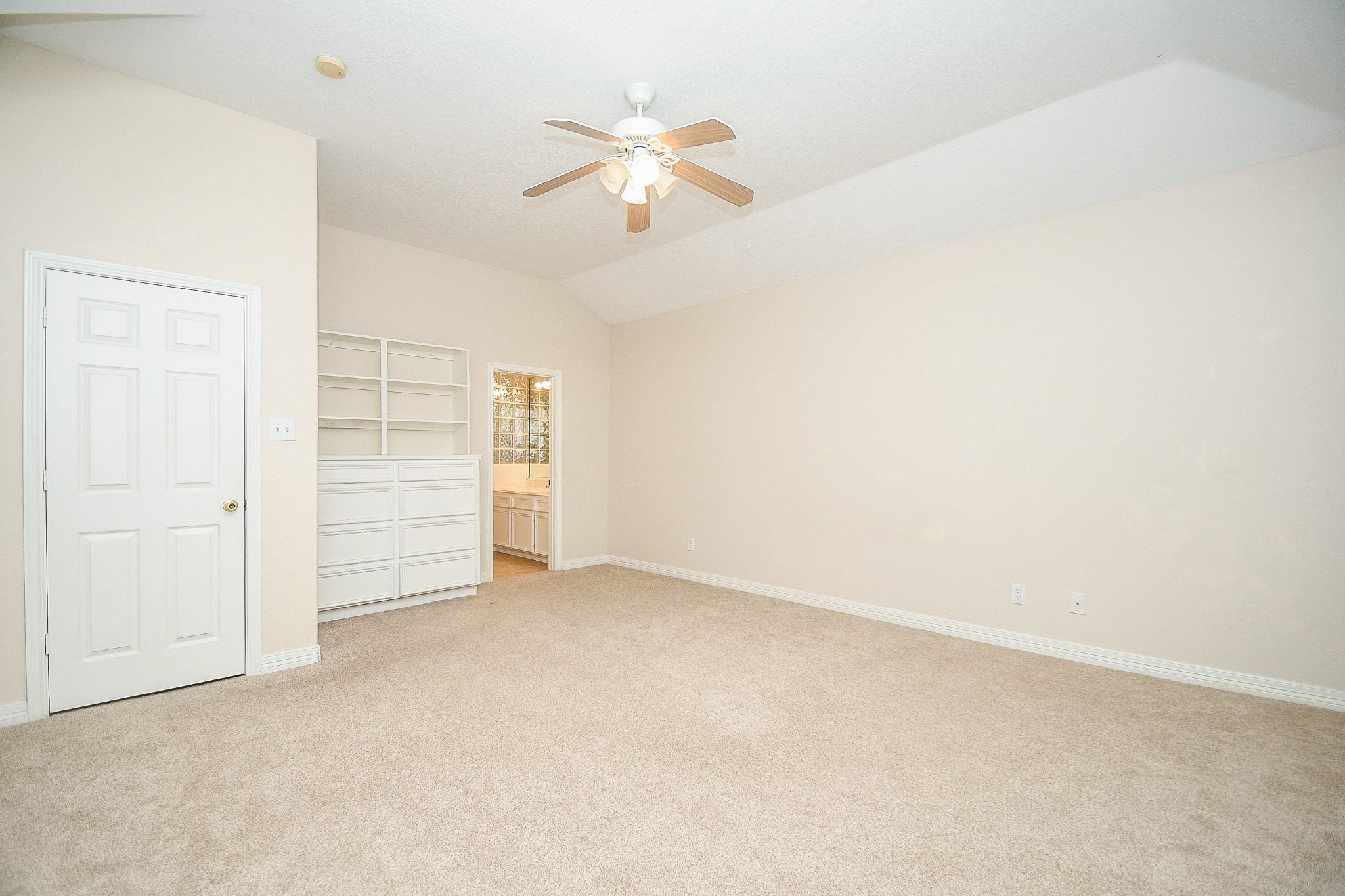 Bright, spacious room with neutral walls and carpet, featuring a ceiling fan. Includes built-in shelves and a view into a bathroom, offering convenience and ample storage.