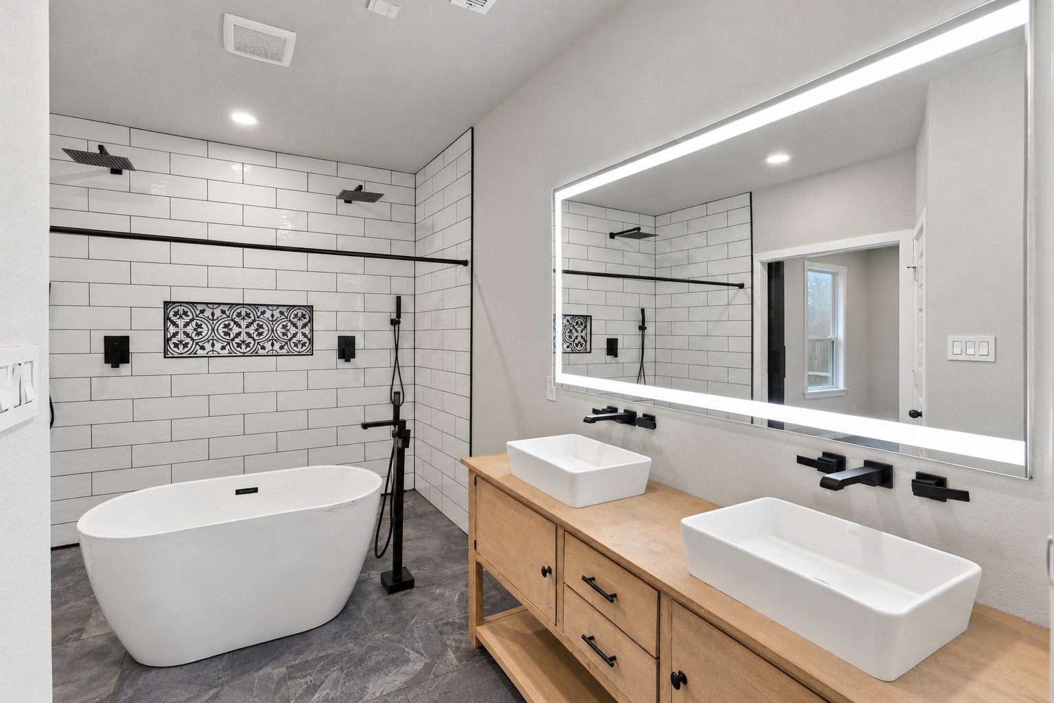 Double vessel sinks, a wood vanity, illuminated mirror, and matte-black fixtures complete the primary bath.