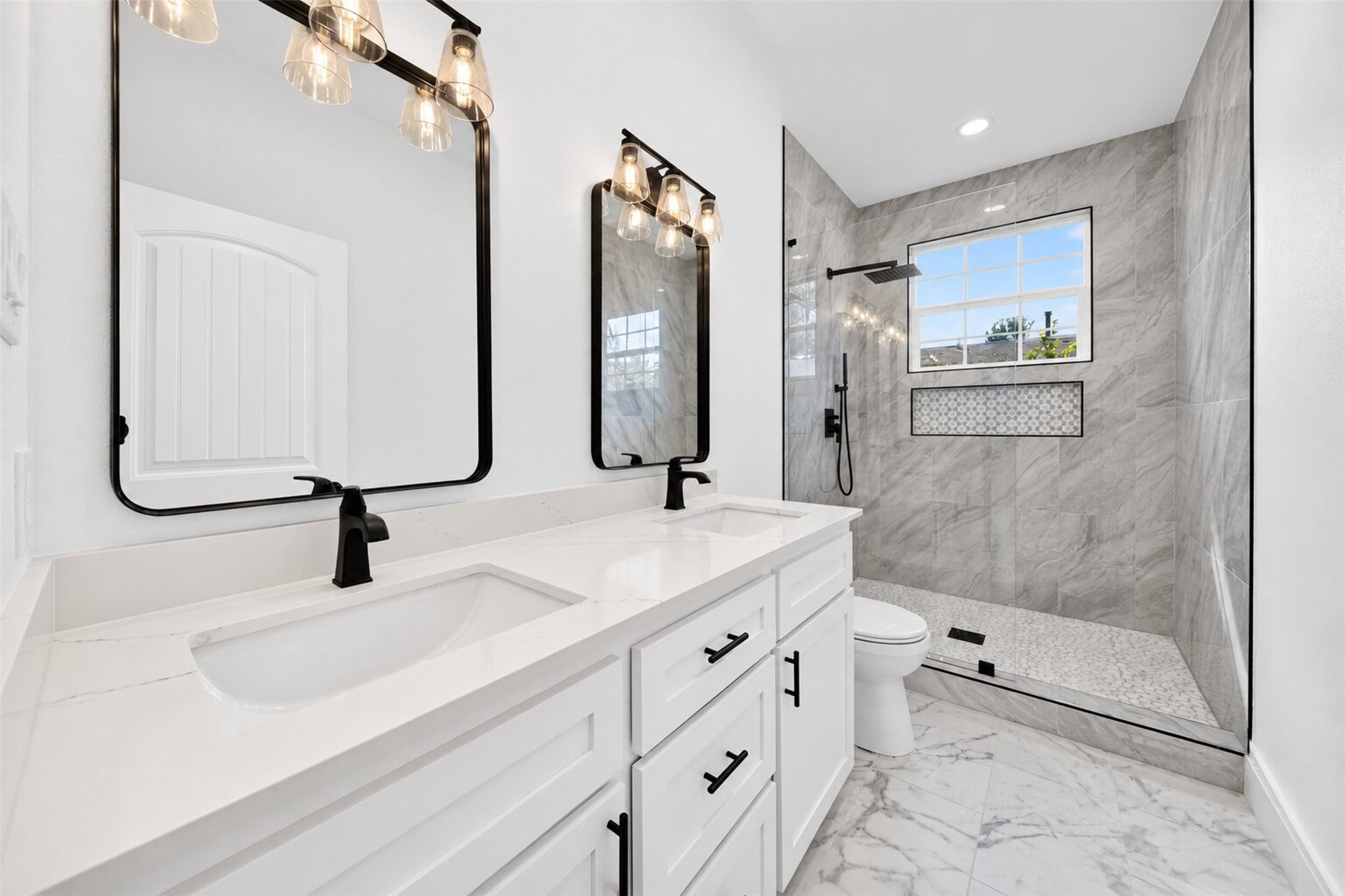 The contemporary bathroom boasts a stylish design, featuring a pristine white vanity, chic black fixtures, and a bathtub adorned with sophisticated gray tile surround. Natural light gently streams through a window, contributing to the clean and luminous ambiance.