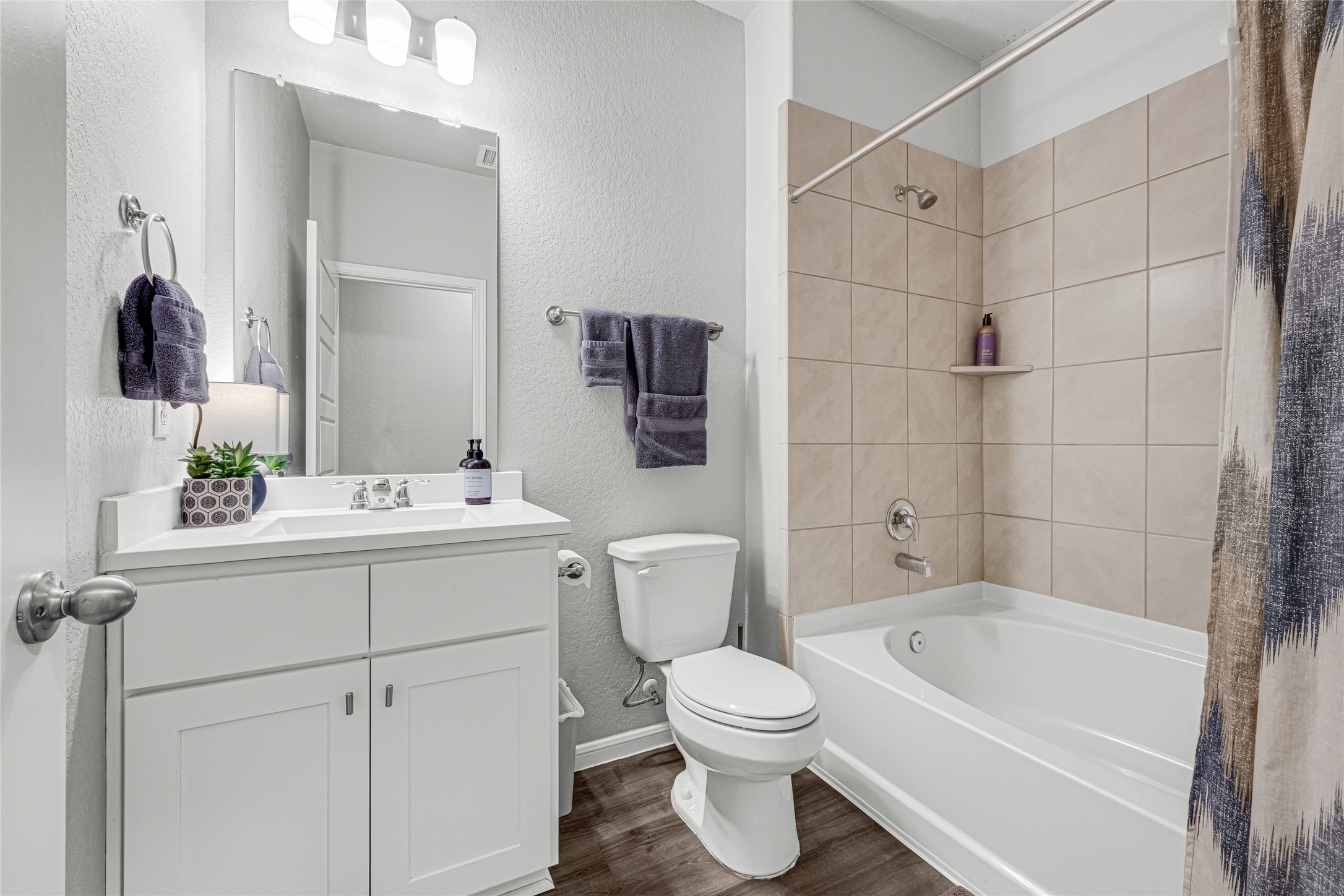 This bathroom features a modern design with a white vanity, large mirror, and sleek lighting with the vinyl plank flooring carrying through. It includes a bathtub with beige tile surround, an toilet, and stylish fixtures, creating a clean and inviting space.