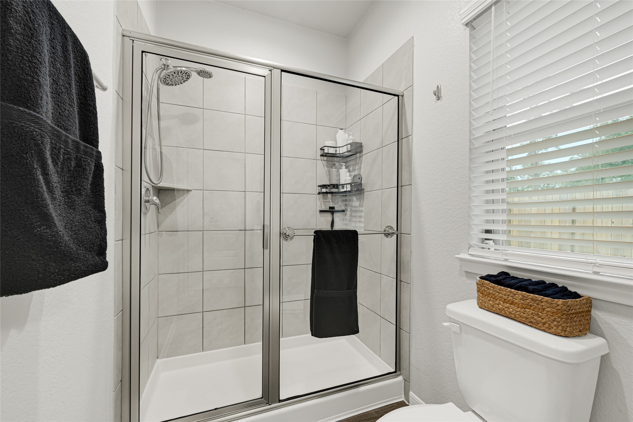 This bathroom features an up graded modern glass-enclosed oversized shower with tiled walls, a window with 2 inch faux wood blinds for natural light, and a neatly organized upgraded elongated commode.  This Primary bathroom offers a spacious interior and plenty of room for your storage needs.