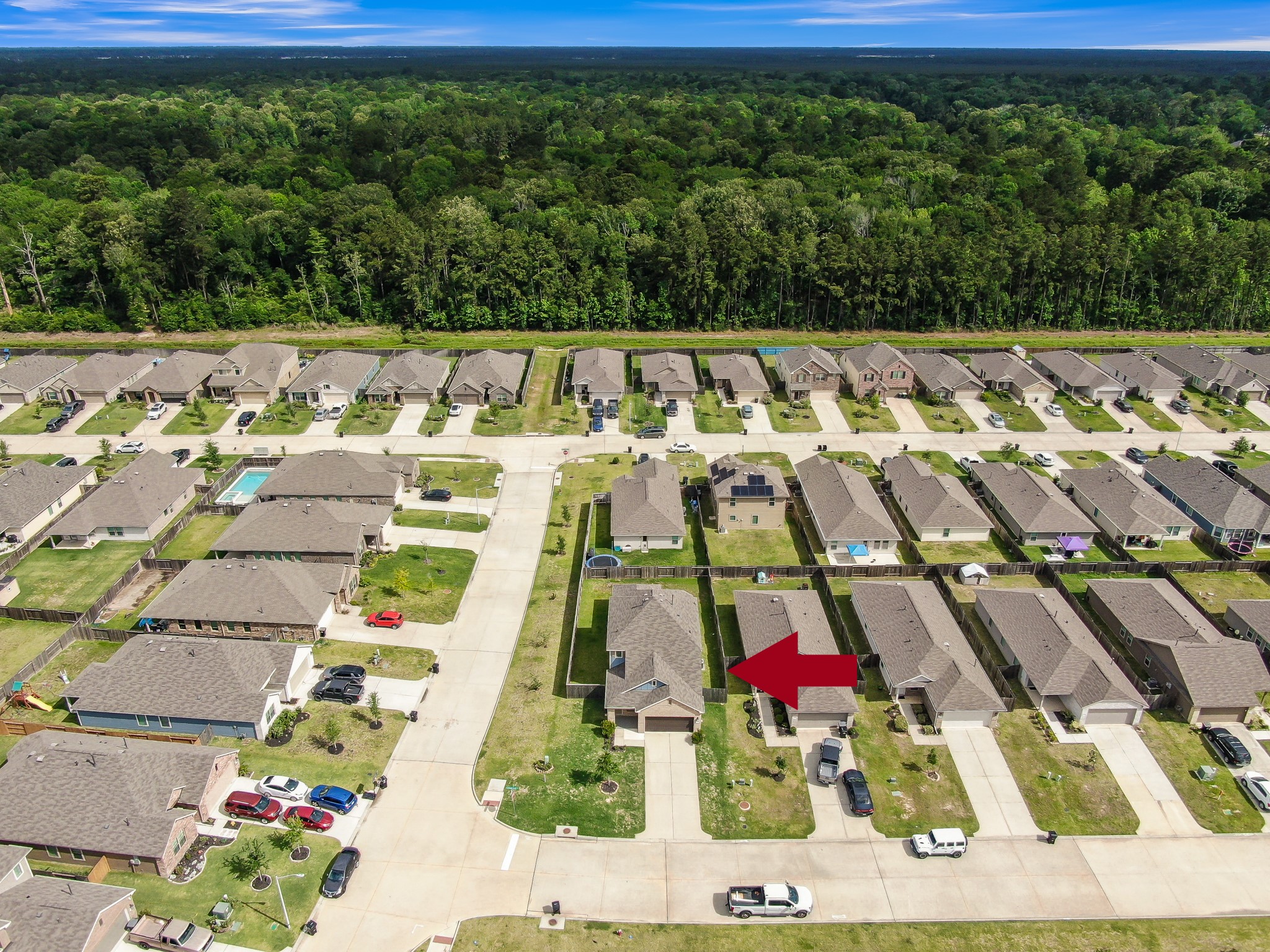 This aerial photo showcases a suburban neighborhood with rows of single-family homes, each with driveways and small front yards. The area is surrounded by lush greenery, providing a sense of tranquility. An arrow points to a specific house, indicating its location within the community.