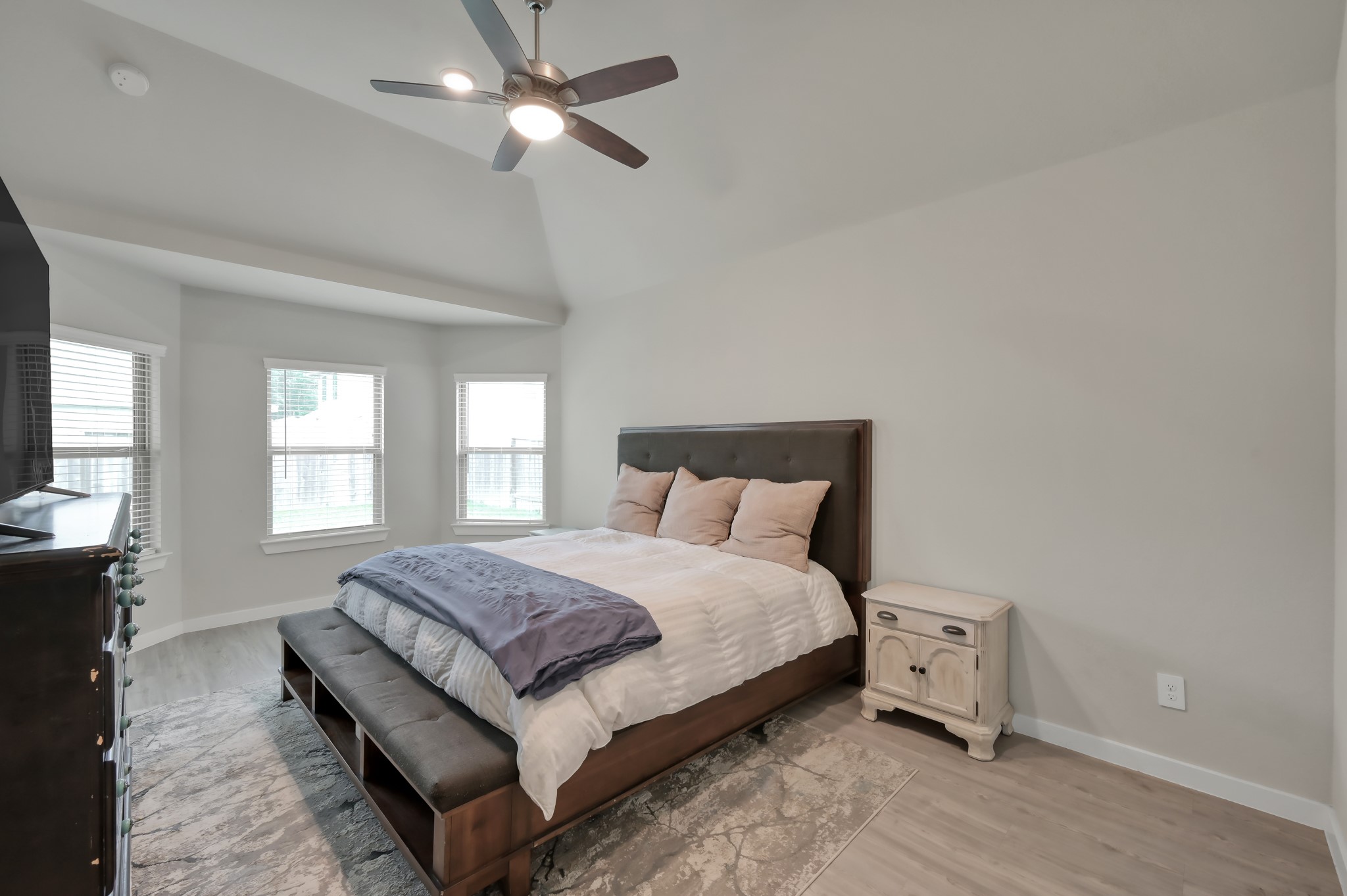 This bedroom features a cozy setup with a large bed, a ceiling fan, and ample natural light from multiple windows. It has a neutral color palette and includes a nightstand and dresser, creating a comfortable and inviting space.
