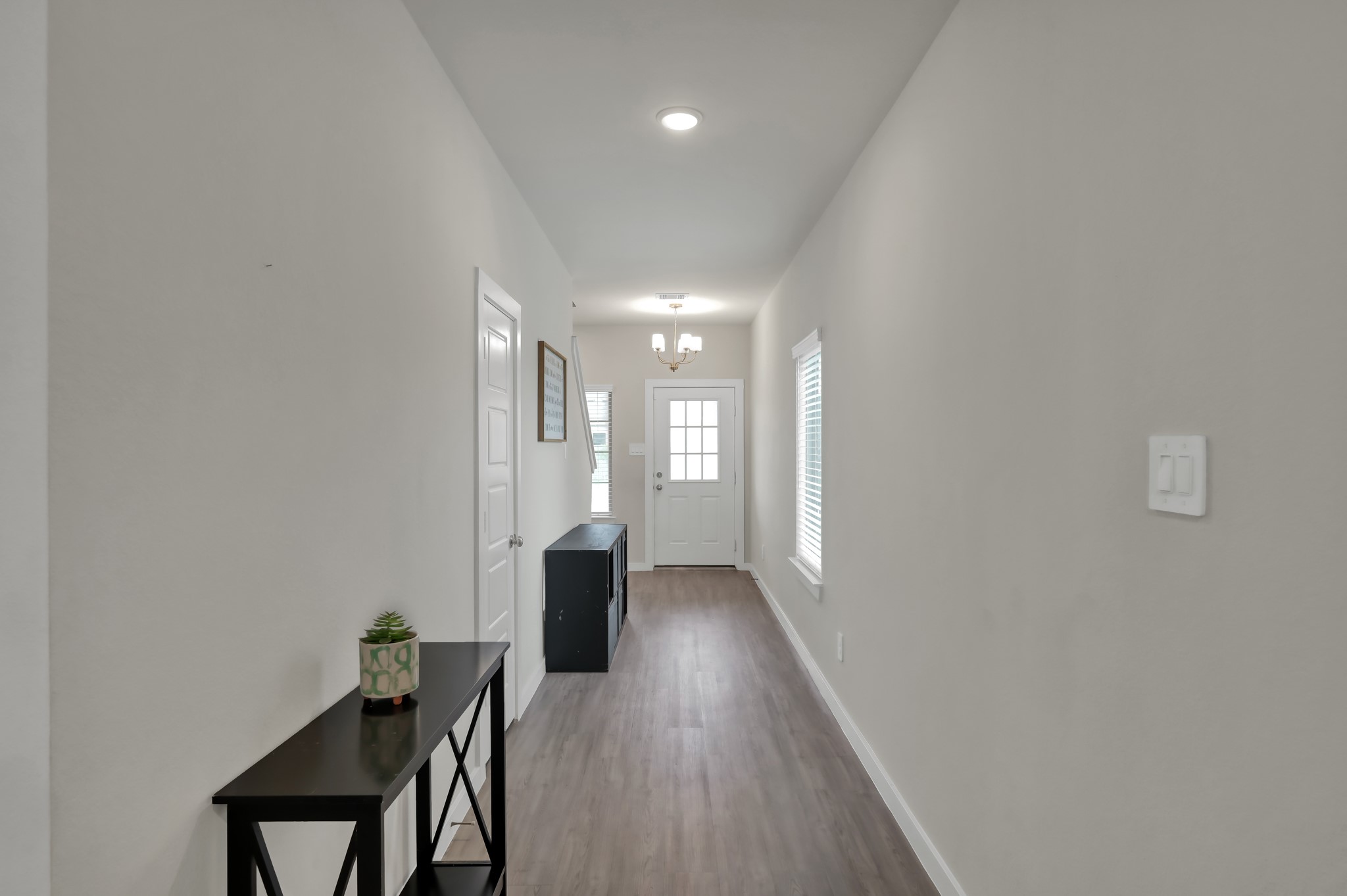 Bright and inviting hallway with neutral walls and wood flooring. Features include a modern light fixture, a side table with decor, a window for natural light, and a door leading to other areas of the home.