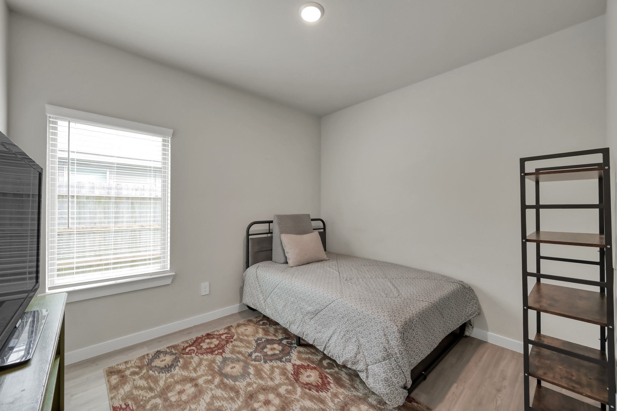 This cozy bedroom features a single bed, a window with blinds for natural light, a patterned area rug, and a minimalist metal shelf. The neutral wall and flooring tones create a calm atmosphere.