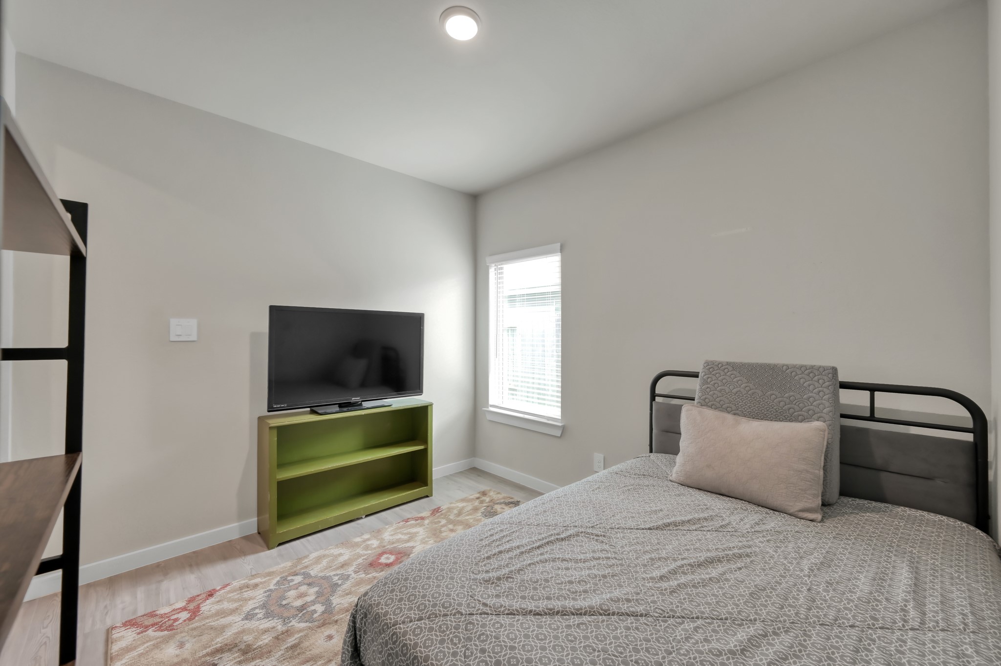 This is a cozy, well-lit bedroom featuring a single bed, a TV on a small green stand, and a window with blinds. The neutral decor and simple design make it inviting and functional.
