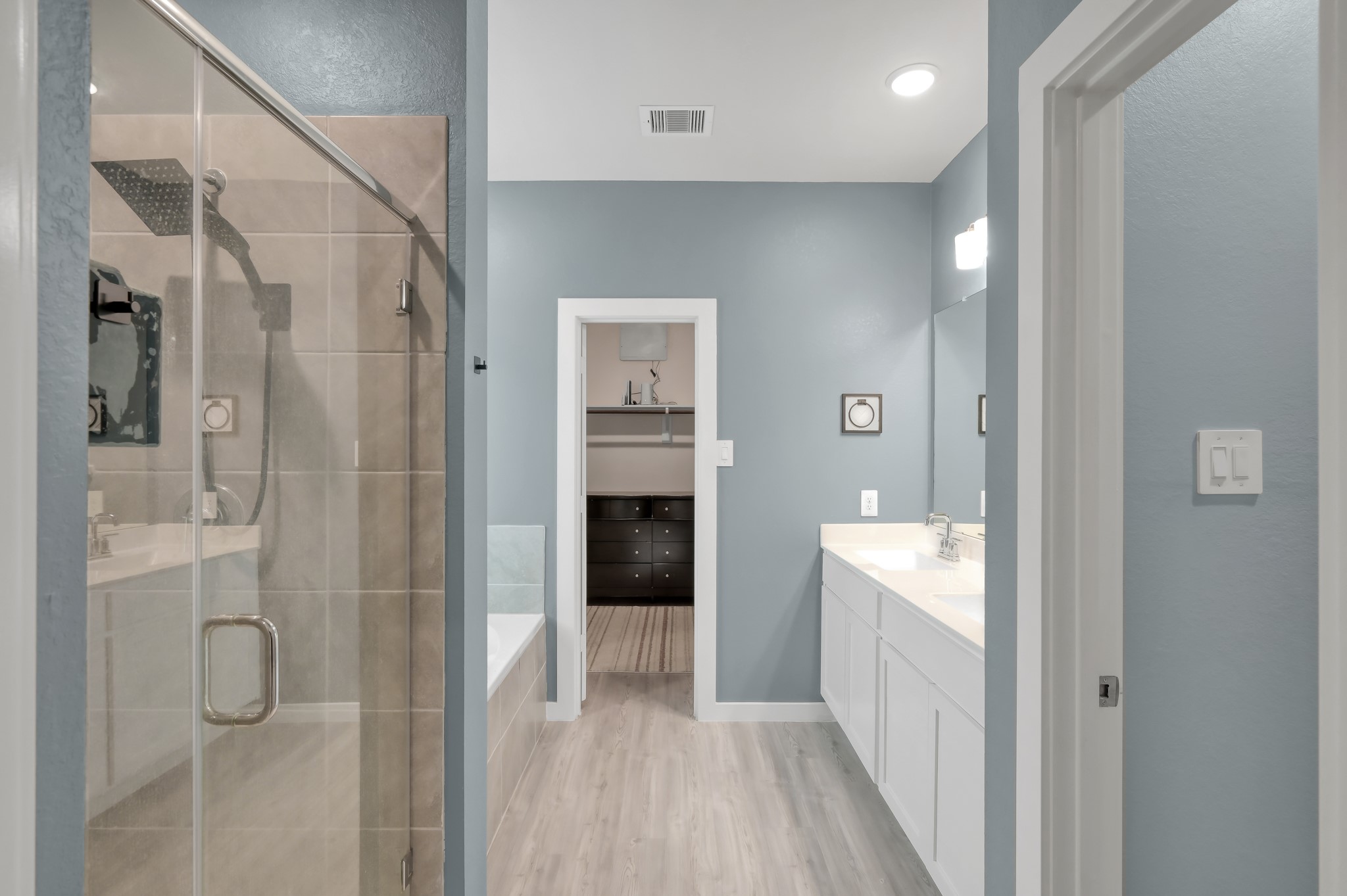 This modern bathroom features a glass-enclosed shower, a separate bathtub, dual sinks with a large mirror, soft blue walls, and light wood flooring, creating a bright and spacious feel.