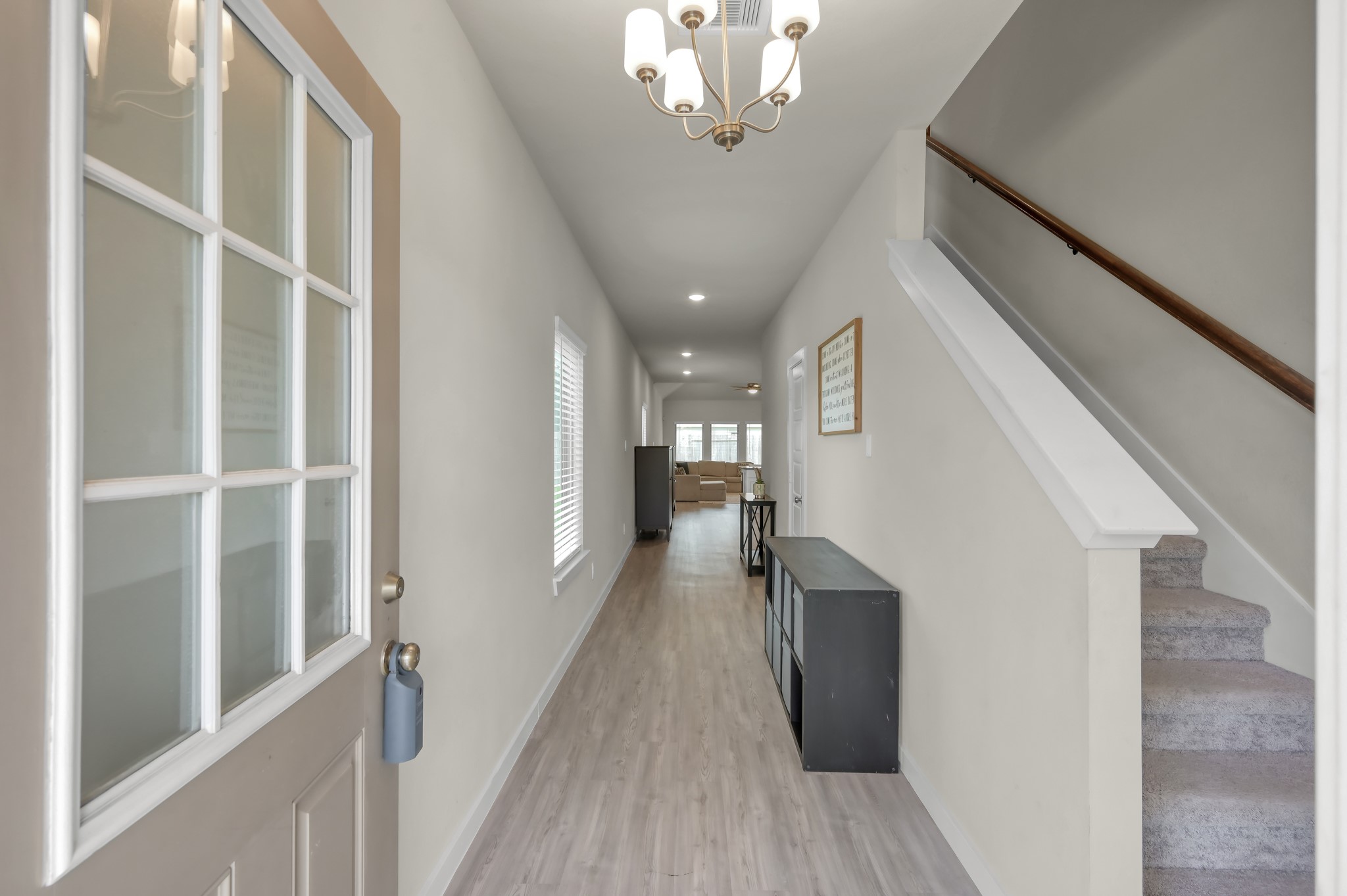 Bright entryway with light wood flooring, modern chandelier, and a staircase leading upstairs. The hallway opens into a spacious living area, offering a welcoming first impression.
