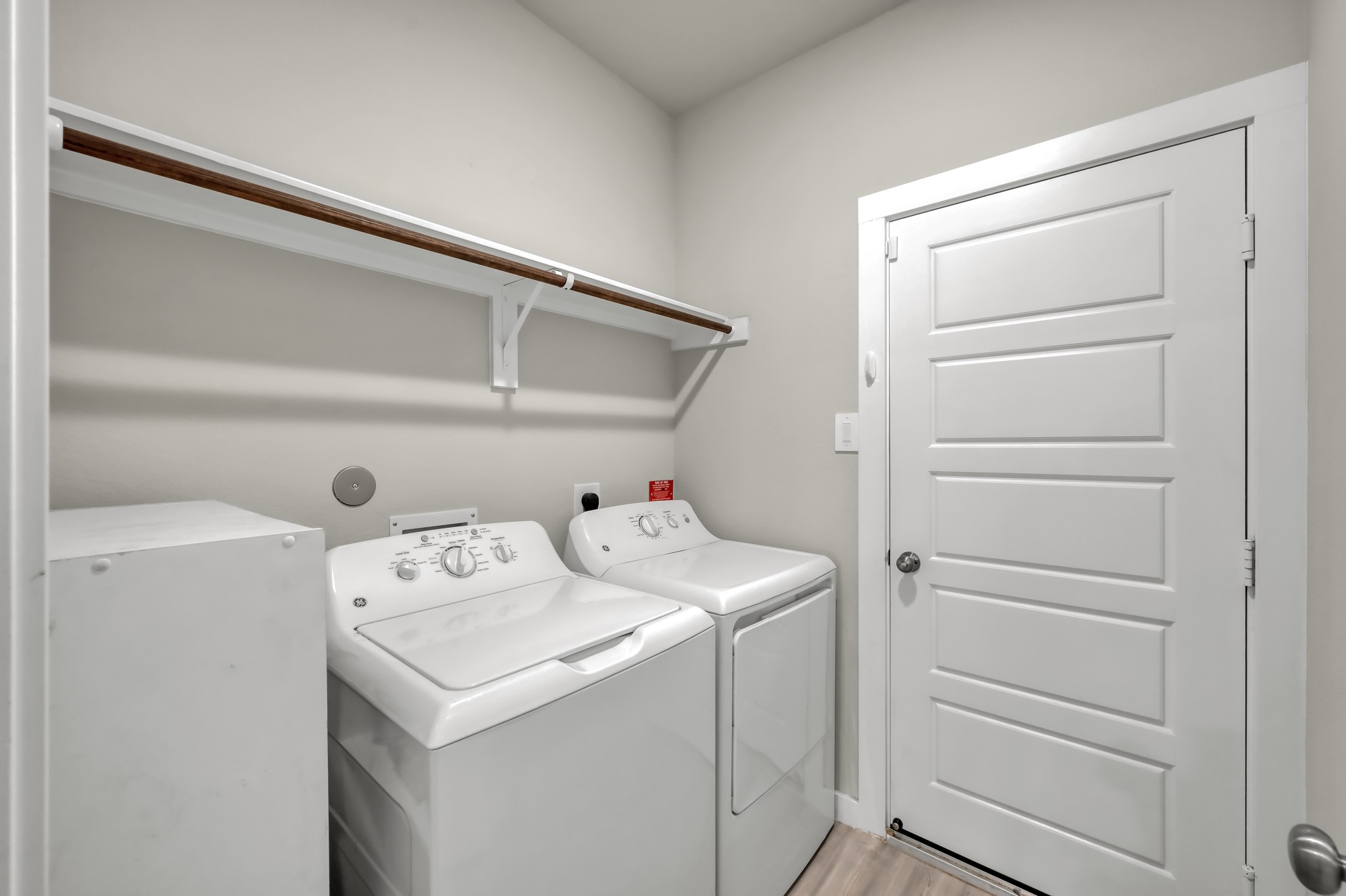Compact laundry room with washer and dryer, featuring a shelf for storage and a white door. Neutral wall color and light wood flooring create a clean, functional space.