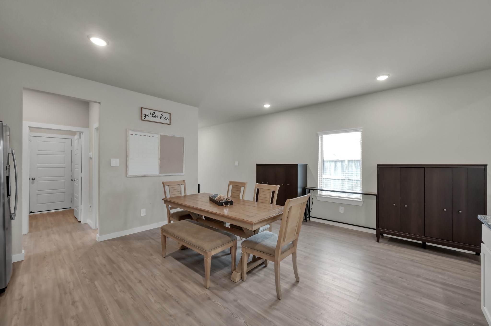 This bright, open dining area features light wood flooring, recessed lighting, and neutral walls. It includes a wooden dining set and ample storage with two large cabinets. The space is adjacent to the kitchen and hallway, offering a welcoming atmosphere.