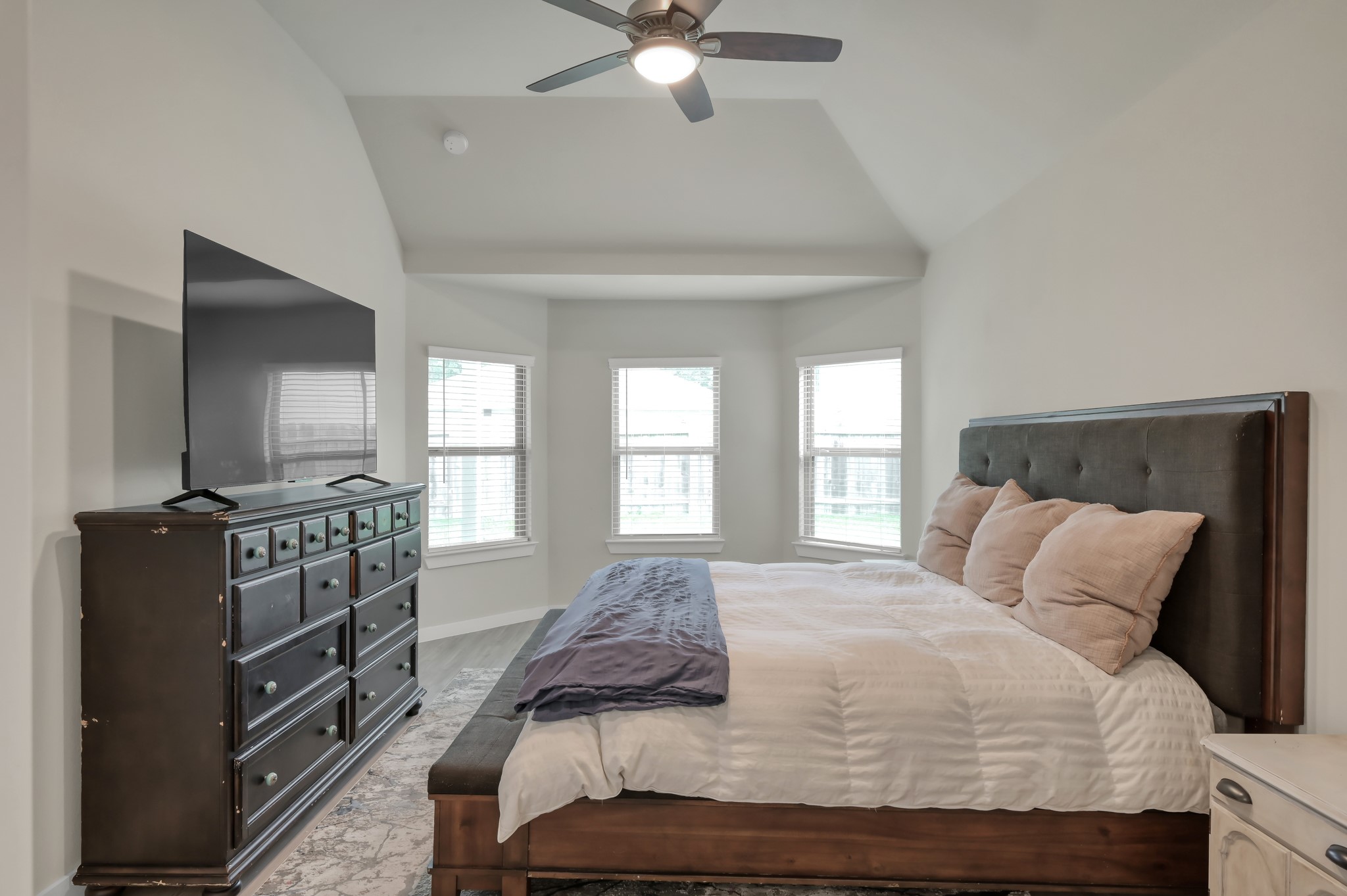 This bedroom is spacious and bright, featuring large windows, a high ceiling with a ceiling fan, and a modern design. It includes a comfortable bed and a large dresser with a mounted TV, offering a cozy yet elegant atmosphere.