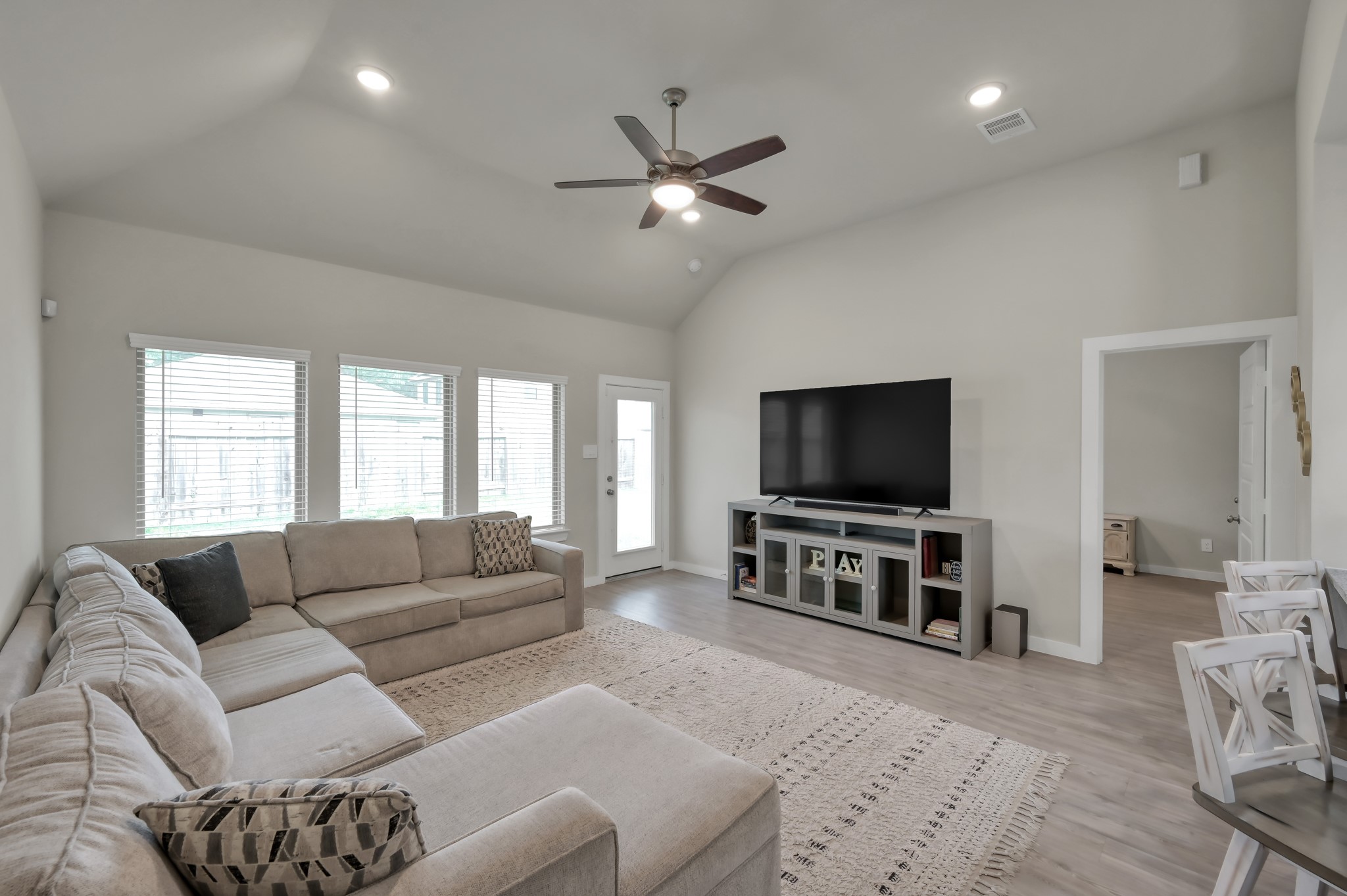 Bright and spacious living room with vaulted ceilings, featuring a large sectional sofa, hardwood floors, and ample natural light from multiple windows. A ceiling fan and modern entertainment setup enhance comfort and functionality.