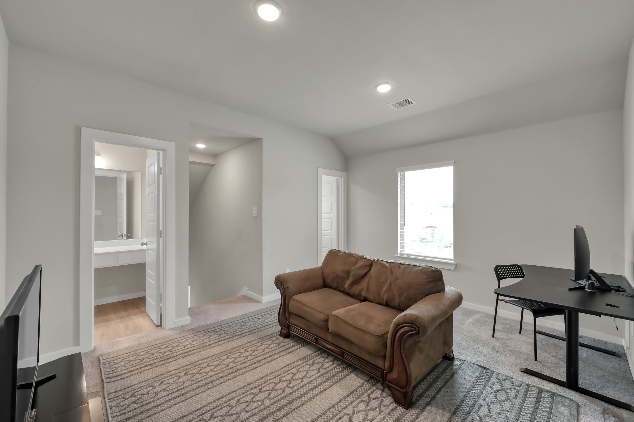 This room offers a cozy living space with a brown sofa, a modern desk setup, and a mounted TV. It features neutral walls, carpeted floors, and ample natural light from a window. A doorway leads to a bathroom, enhancing functionality. Ideal for a home office or relaxation area.