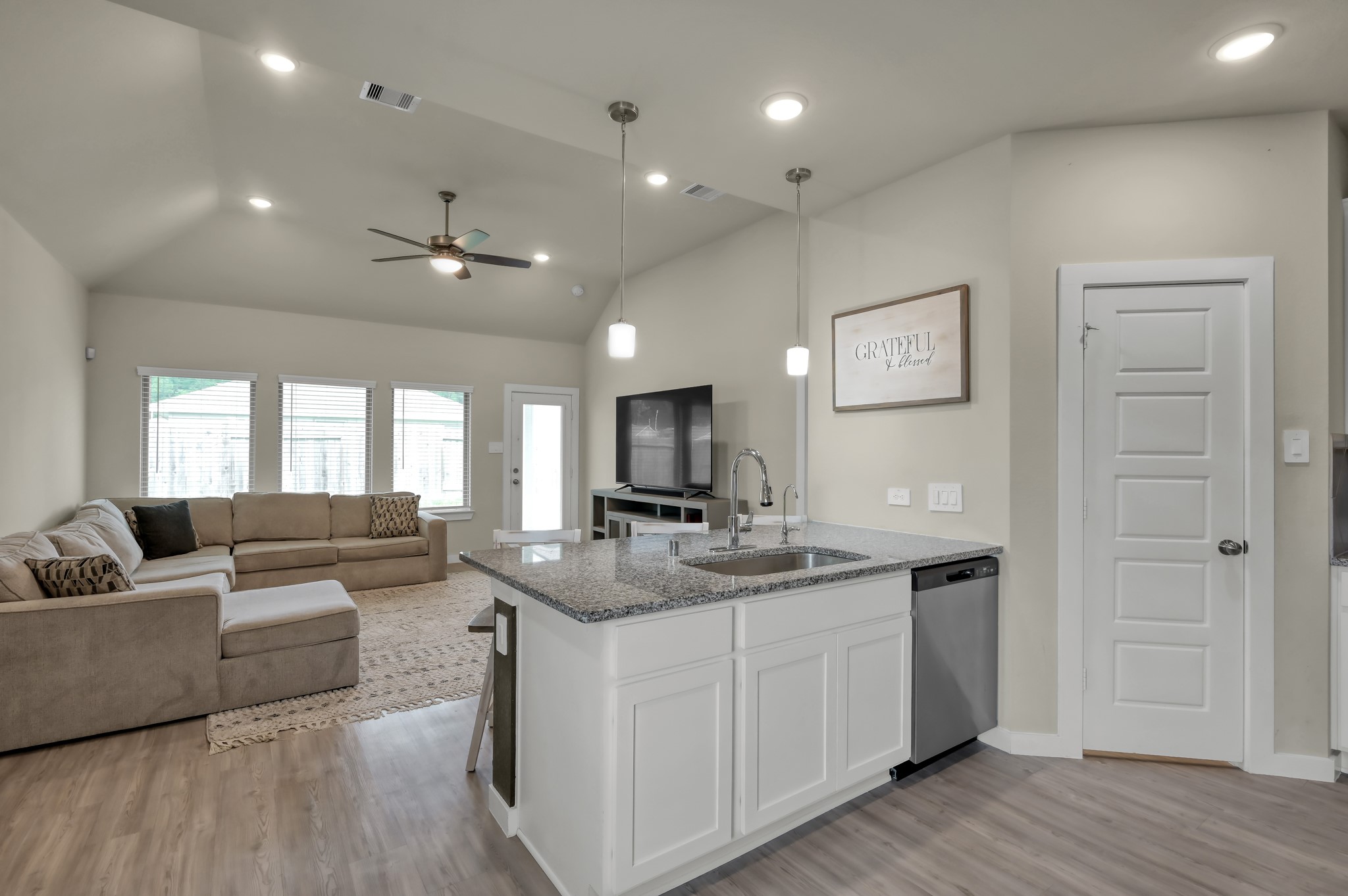 This modern, open-concept living space features a cozy sectional sofa, large windows for natural light, and a central kitchen island with granite countertops. The light wood flooring and neutral walls create a warm, inviting atmosphere.