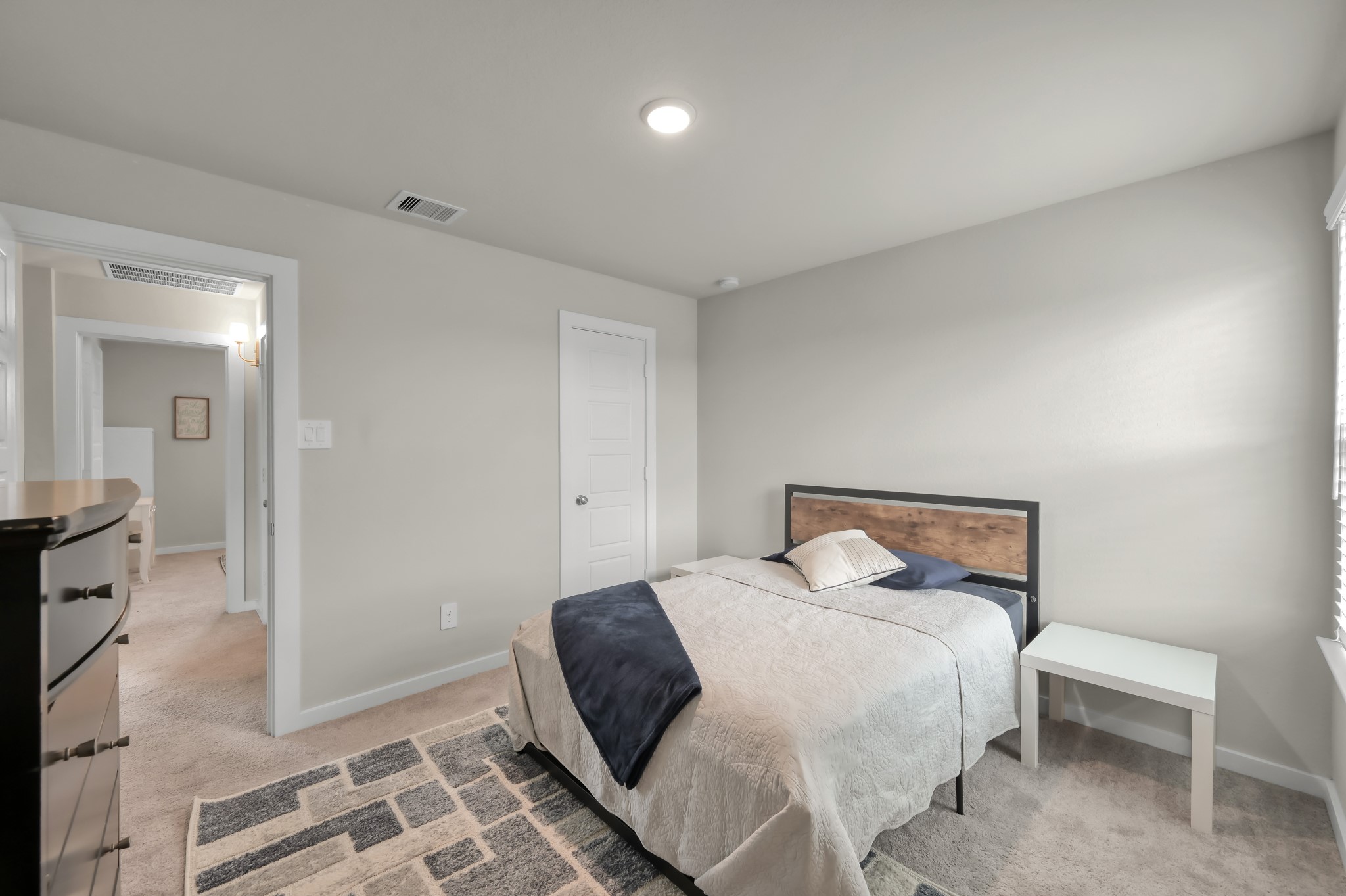 A cozy bedroom with neutral tones, featuring a bed with a wooden headboard, a side table, and a dresser. The room has carpeted flooring and is well-lit, providing a warm and inviting atmosphere.