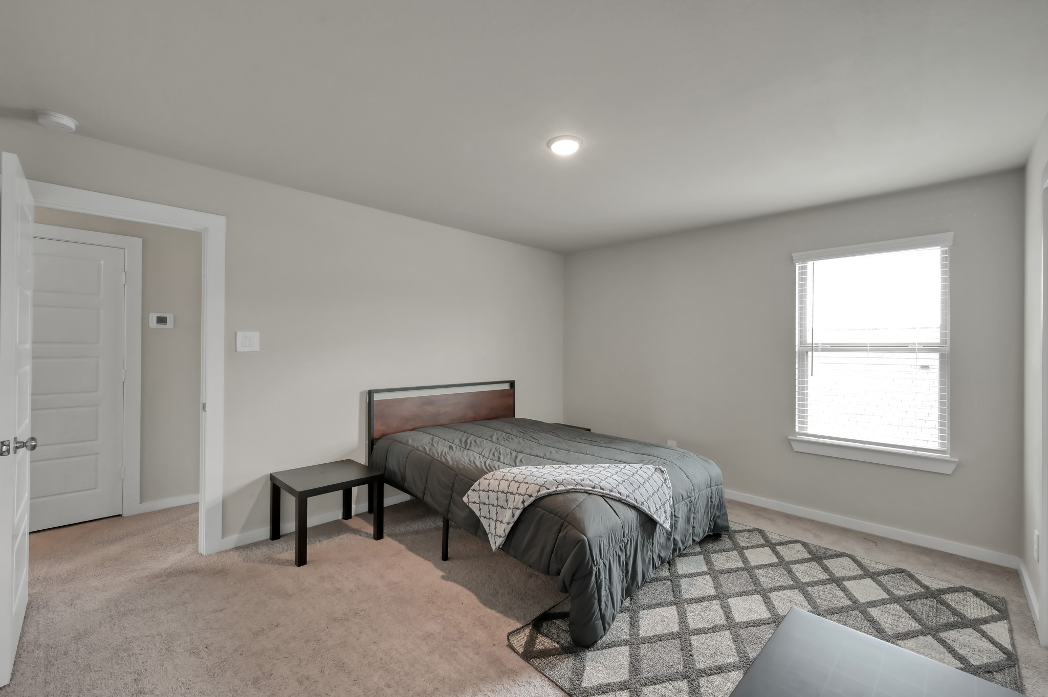 This cozy bedroom features neutral walls, a carpeted floor, and ample natural light from a single window. It includes a bed with a simple headboard and a small side table, creating a comfortable, minimalist space.