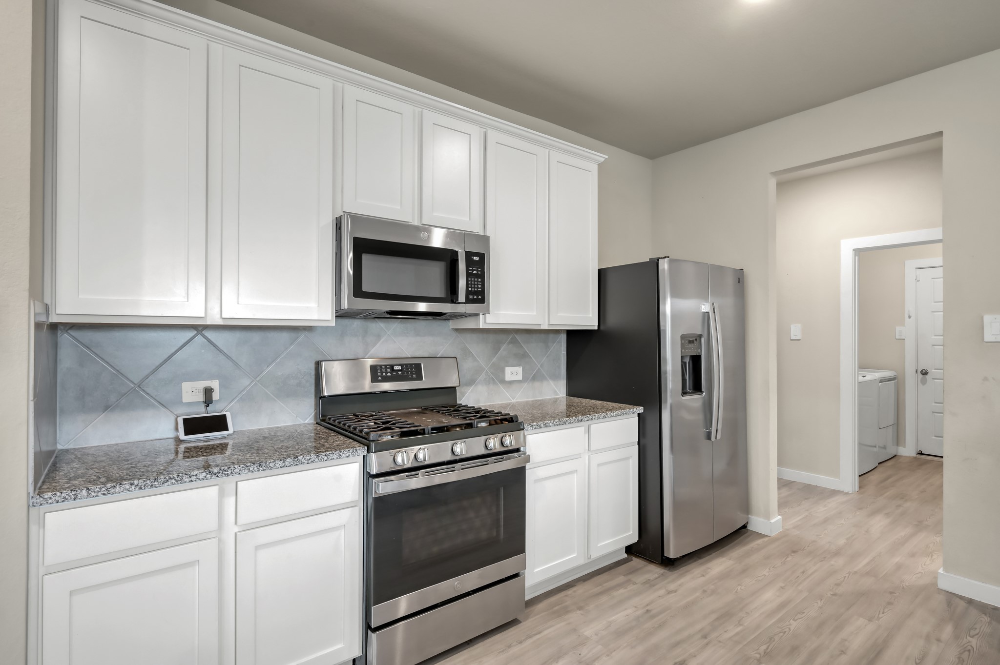 This modern kitchen with sleek stainless steel appliances, including a gas stove and refrigerator. Features white cabinetry, granite countertops, and a stylish tile backsplash. Adjacent to a utility room with washer and dryer.
