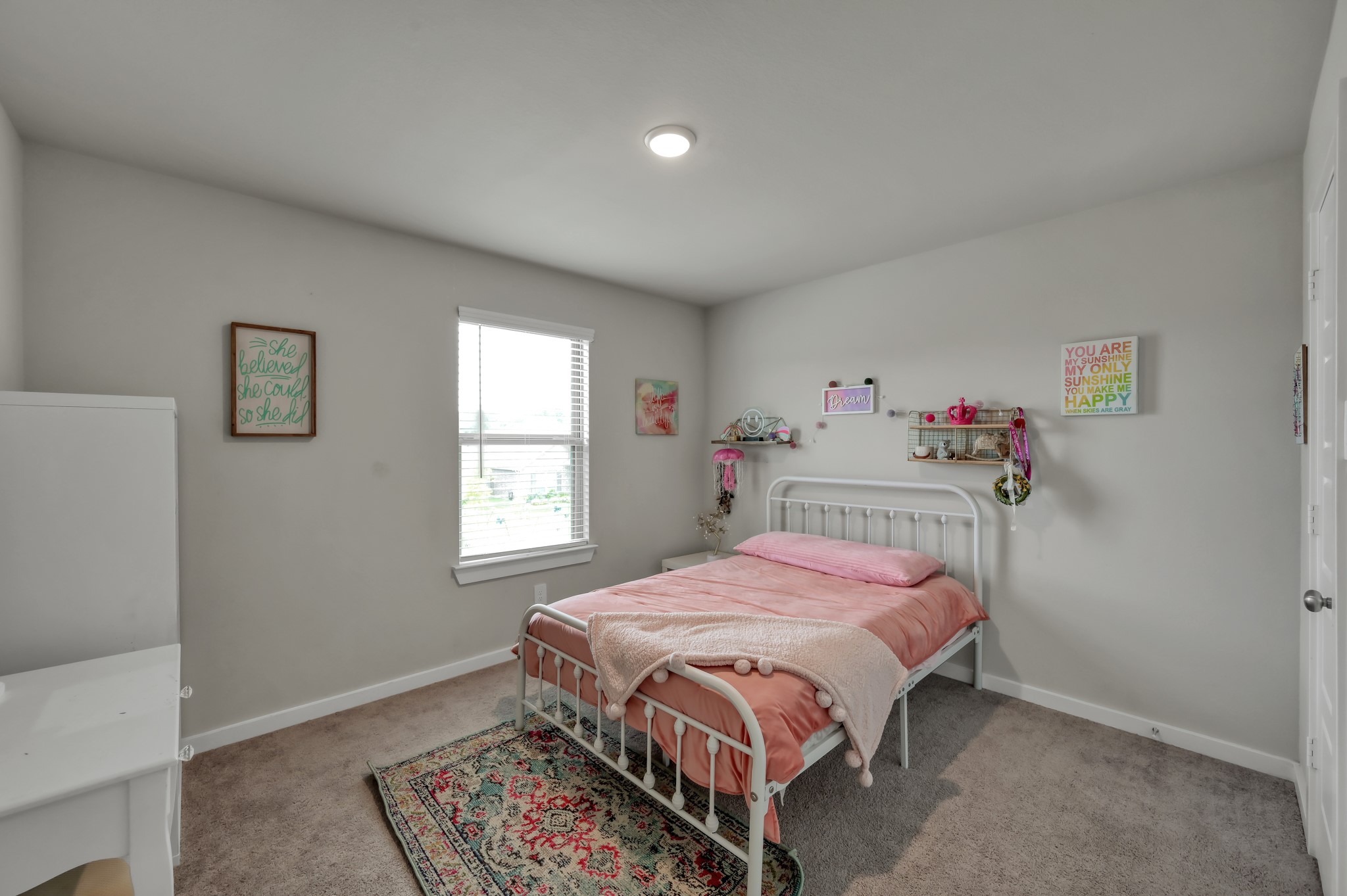 This cozy, well-lit bedroom features soft carpeting, a large window for natural light, and a neatly made bed. The walls are adorned with cheerful decorations, creating a welcoming and comfortable space.