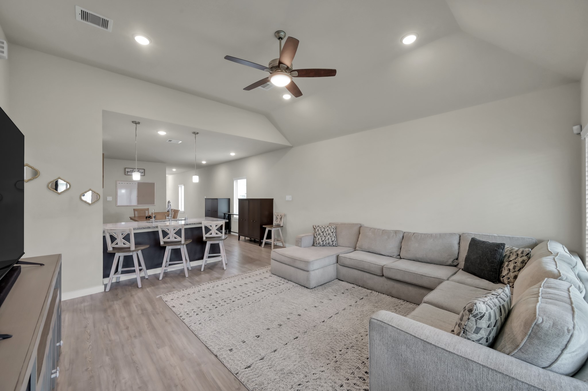 A spacious, open-concept living area featuring a large sectional sofa, modern ceiling fan, and recessed lighting. The room flows seamlessly into the kitchen, which has a breakfast bar with stylish pendant lights and seating. Neutral tones and sleek flooring enhance the contemporary feel.