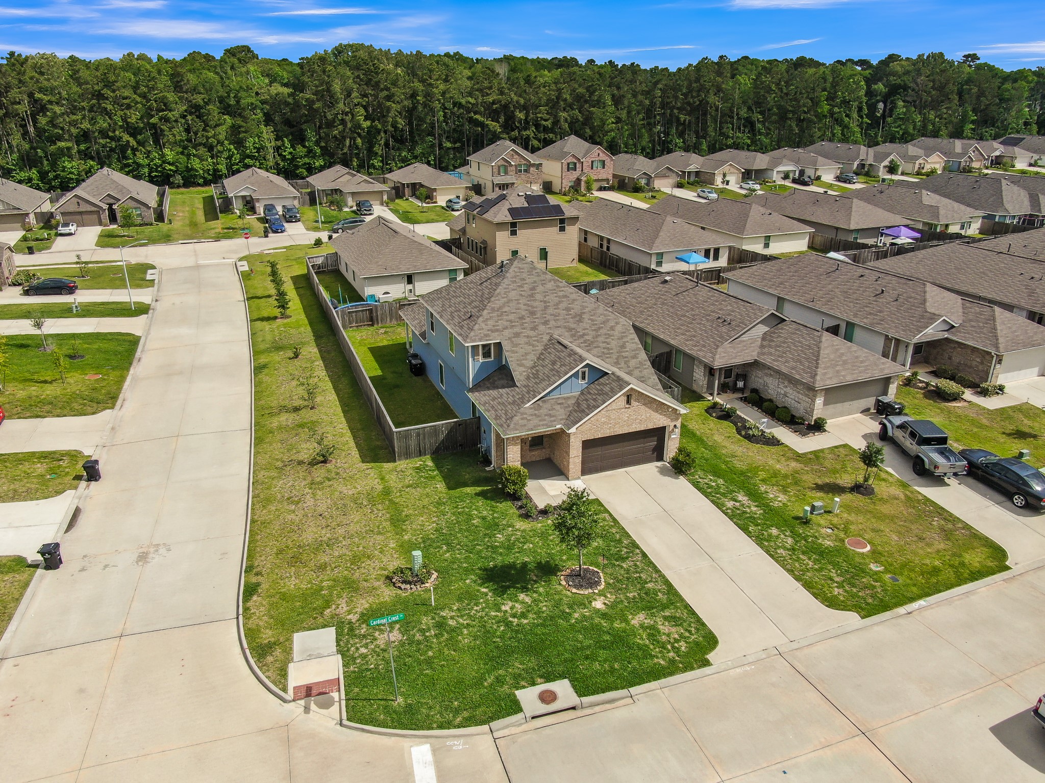 Aerial view of a suburban neighborhood featuring a single-story home with a two-car garage and well-maintained lawn. The house is situated on a corner lot with a spacious backyard and privacy fence. The street is lined with similar houses and bordered by a wooded area.
