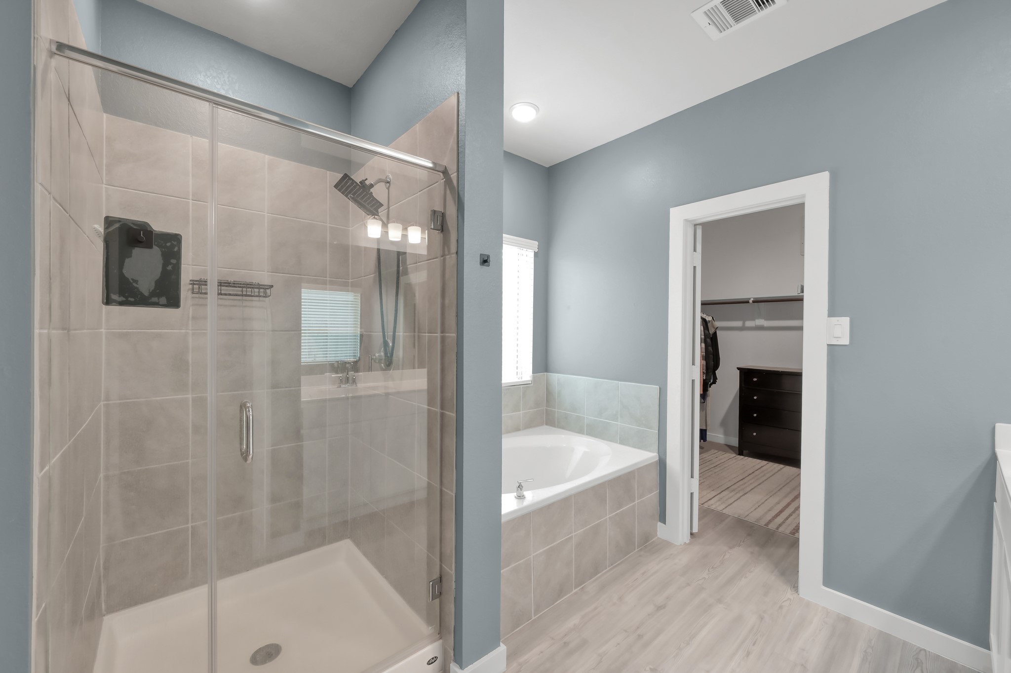 A modern bathroom featuring a glass-enclosed shower, a bathtub, and an adjoining walk-in closet. The room has a clean, contemporary design with light blue walls and ample natural light.