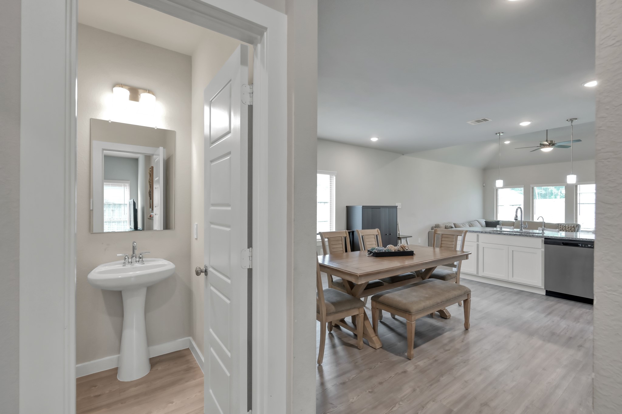 An open-concept living space featuring a dining area with a wooden table set and a modern kitchen with white cabinetry and stainless steel appliances. There's also a glimpse of a small bathroom with a pedestal sink and mirror. The area has light wood flooring and ample natural light from large windows.