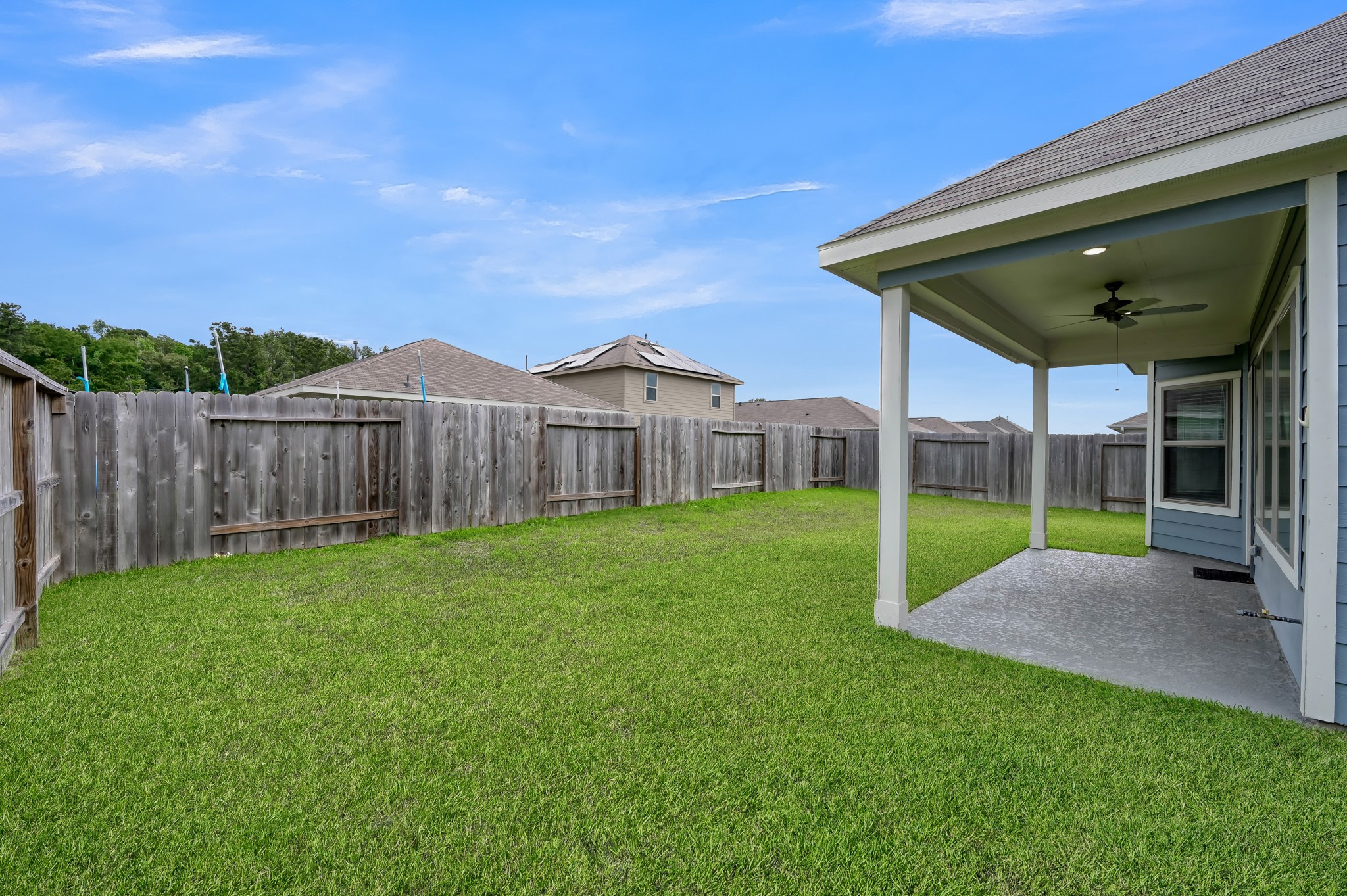 A spacious backyard with a well-maintained lawn, enclosed by a wooden fence for privacy. A covered patio area with a ceiling fan provides a comfortable spot for outdoor relaxation or entertaining.