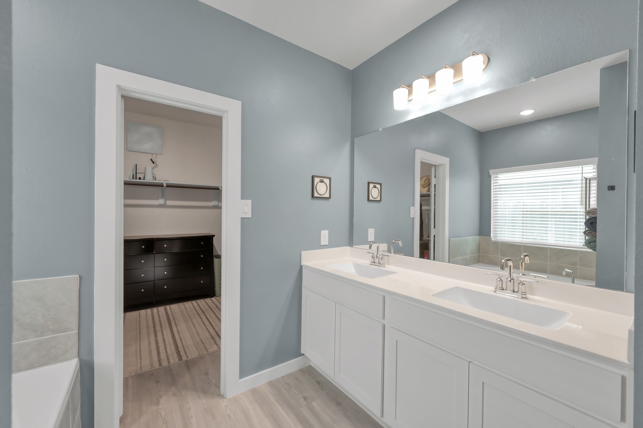 A modern bathroom with a dual-sink vanity, sleek white cabinets, and elegant fixtures. The room features a large mirror, soft blue walls, and ample natural light from a window. A walk-in closet is visible through an open door, offering additional storage space.
