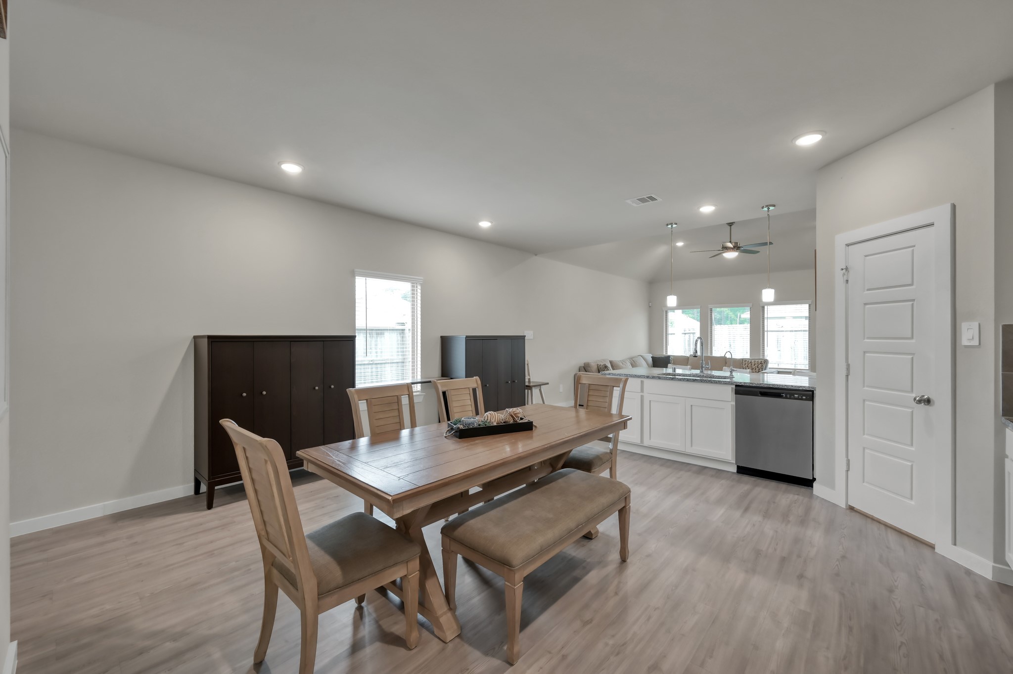 A modern, open-concept dining and kitchen area with a wooden dining set, light wood flooring, and neutral walls. The kitchen features white cabinetry, stainless steel appliances, and pendant lighting over the counter.