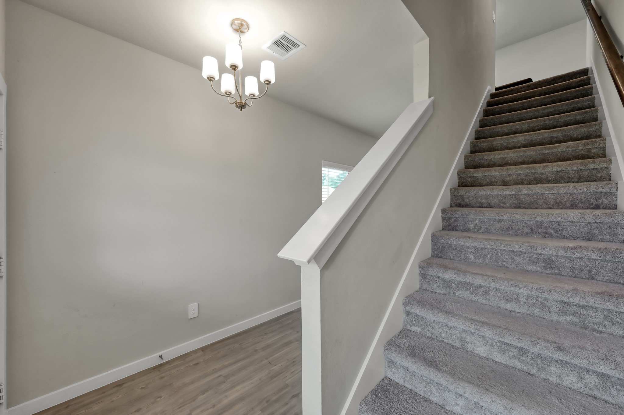 A modern, well-lit entryway with carpeted stairs leading to the upper floor. The space features light wood flooring and a simple chandelier for a welcoming ambiance.