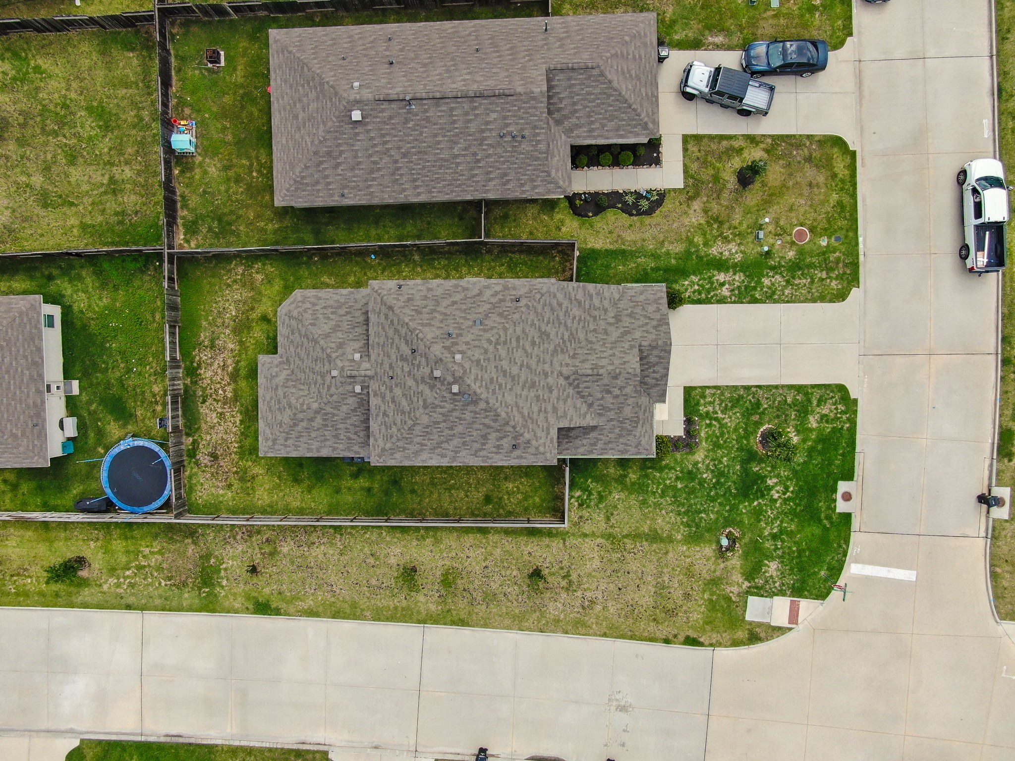 This aerial photo shows a suburban home with a large fenced backyard, a trampoline, and a driveway with parked vehicles. The house is situated on a corner lot in a neighborhood with similar homes.