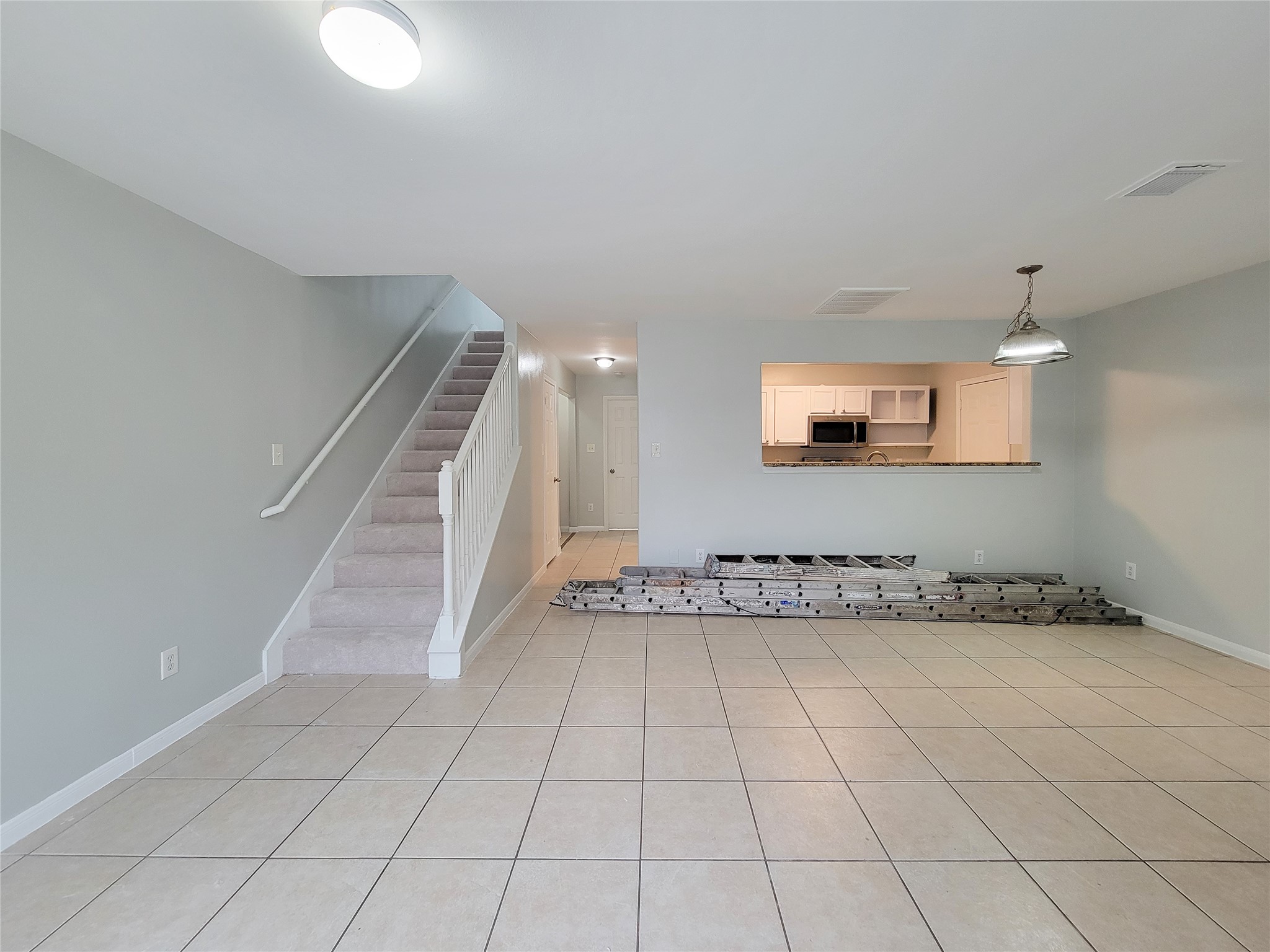 Spacious open-plan living area with neutral tile flooring, leading to a cozy carpeted staircase, and featuring a modern kitchen with a convenient pass-through window.