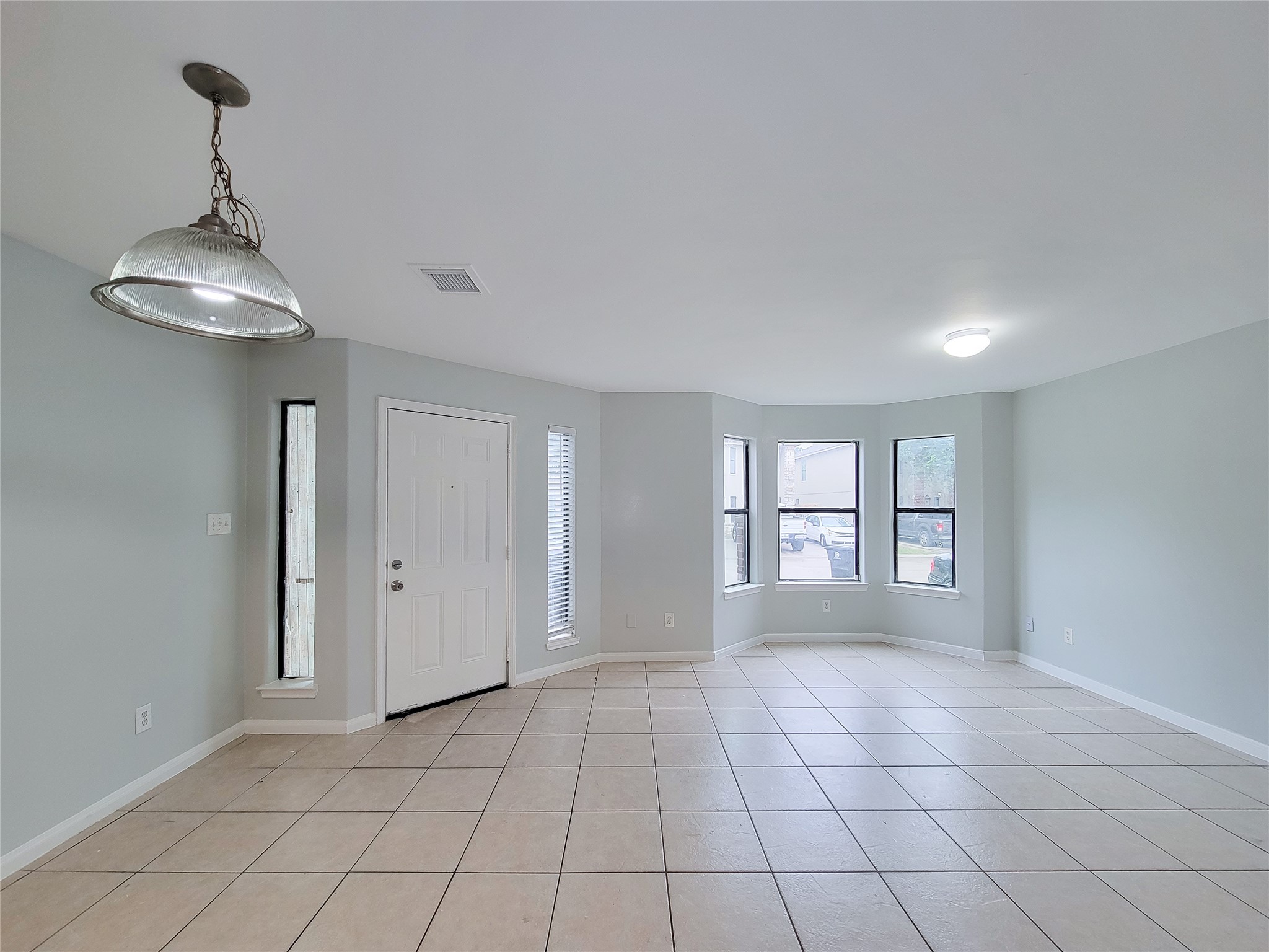 Spacious and bright living area with large windows, neutral tile flooring, and a stylish overhead light fixture, perfect for versatile decorating.