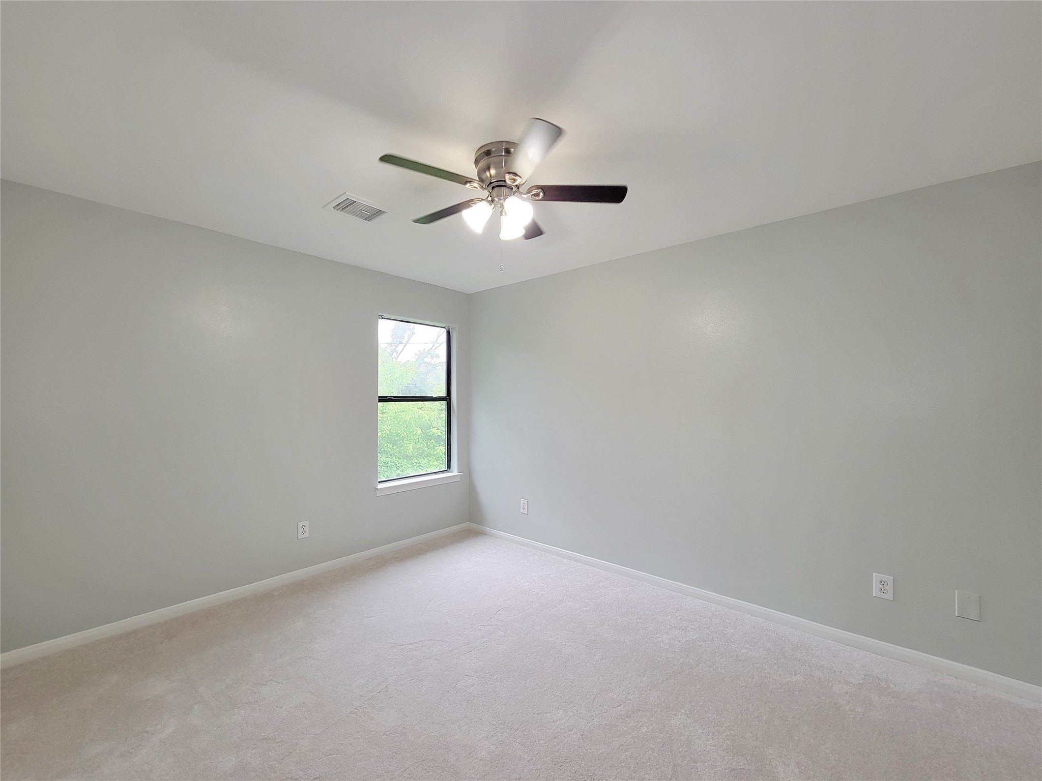 Bright and spacious bedroom featuring soft gray walls, plush carpeting, a large window for natural light, and a modern ceiling fan for comfort.