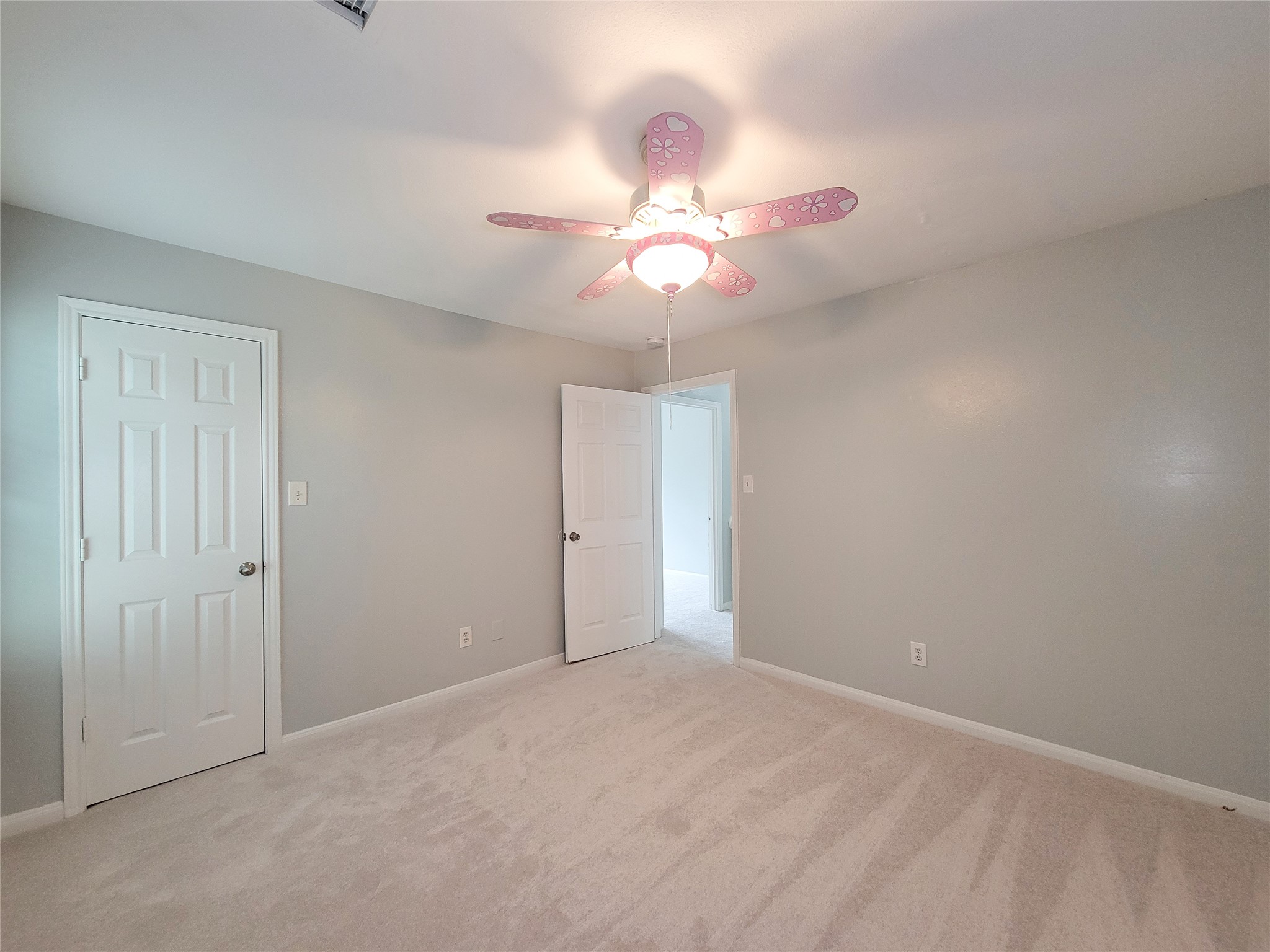 Bright and inviting bedroom featuring soft gray walls, plush carpet, and a playful pink ceiling fan, offering a cozy and cheerful ambiance.