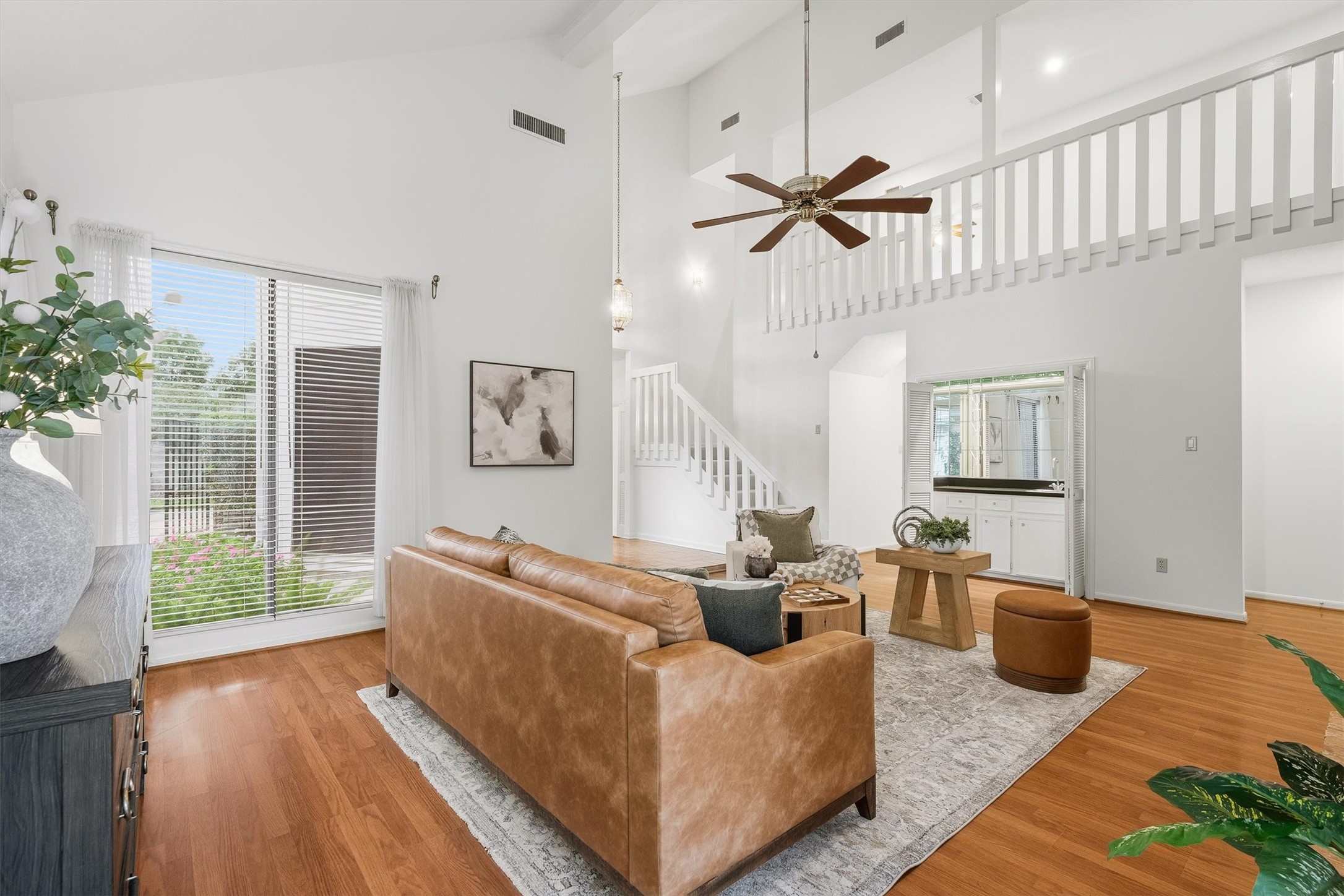 Spacious living room with high ceilings, featuring a cozy leather sofa, elegant hardwood floors, and abundant natural light from large windows. A stylish staircase and modern ceiling fan add to the contemporary charm.