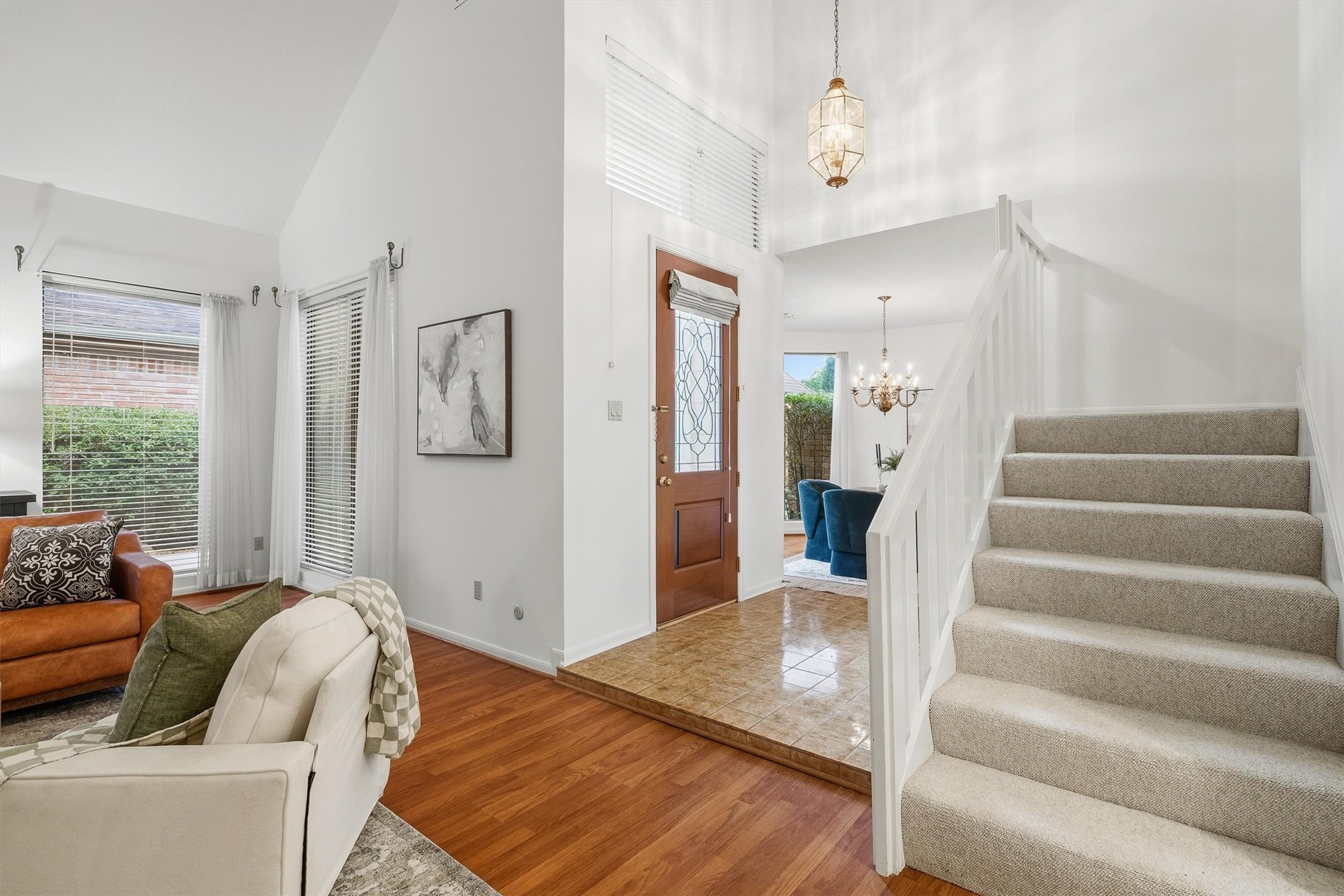 Bright and welcoming entryway with a carpeted staircase, hardwood floors, and a chandelier-lit dining area, leading to a cozy living space with large windows.