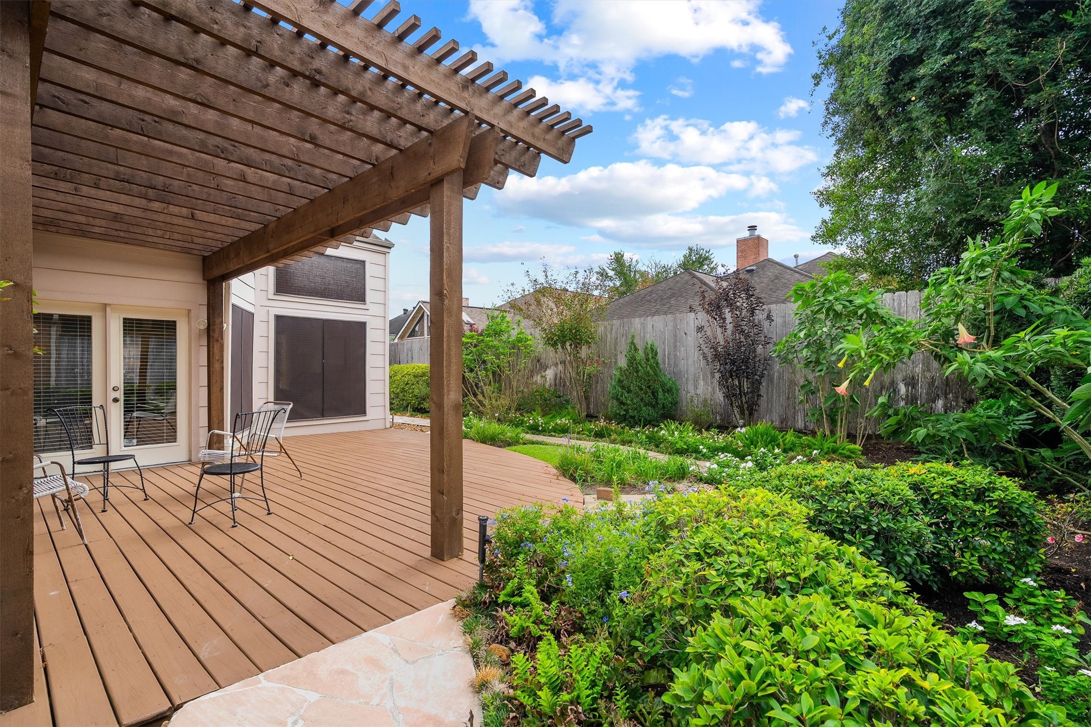 Experience serene outdoor living with this charming wooden deck featuring a pergola, surrounded by lush greenery and vibrant gardens.