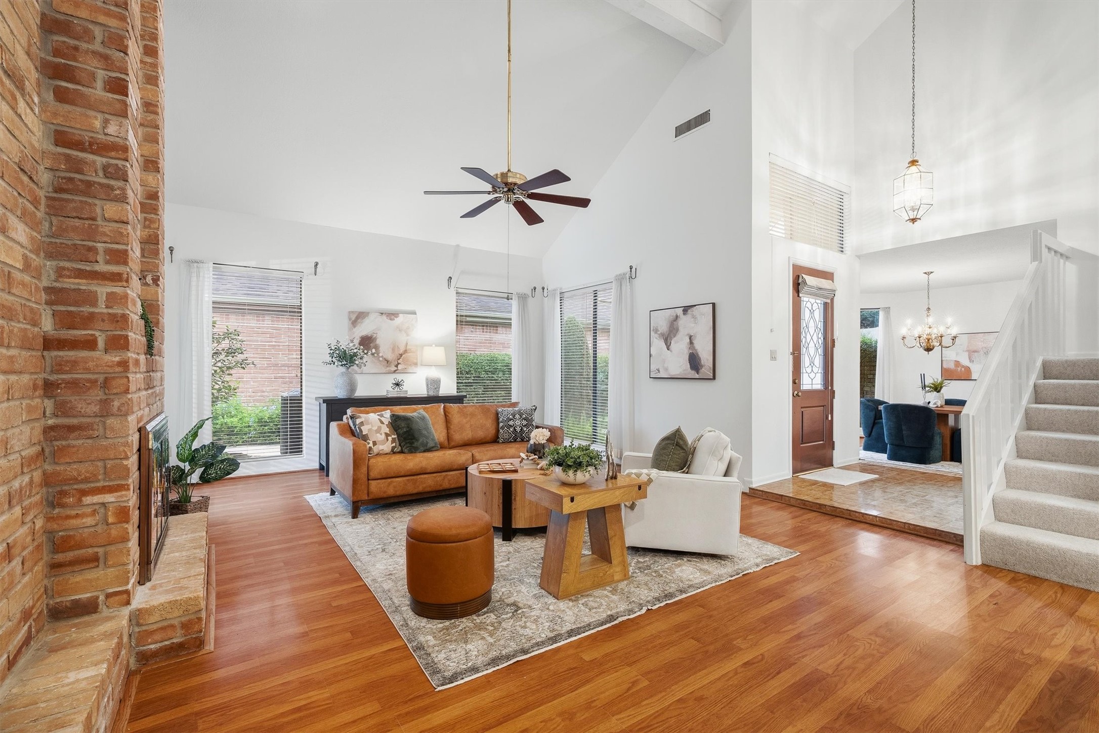 Bright and inviting living space featuring a vaulted ceiling, cozy brick fireplace, and elegant hardwood floors, seamlessly flowing into the dining area.