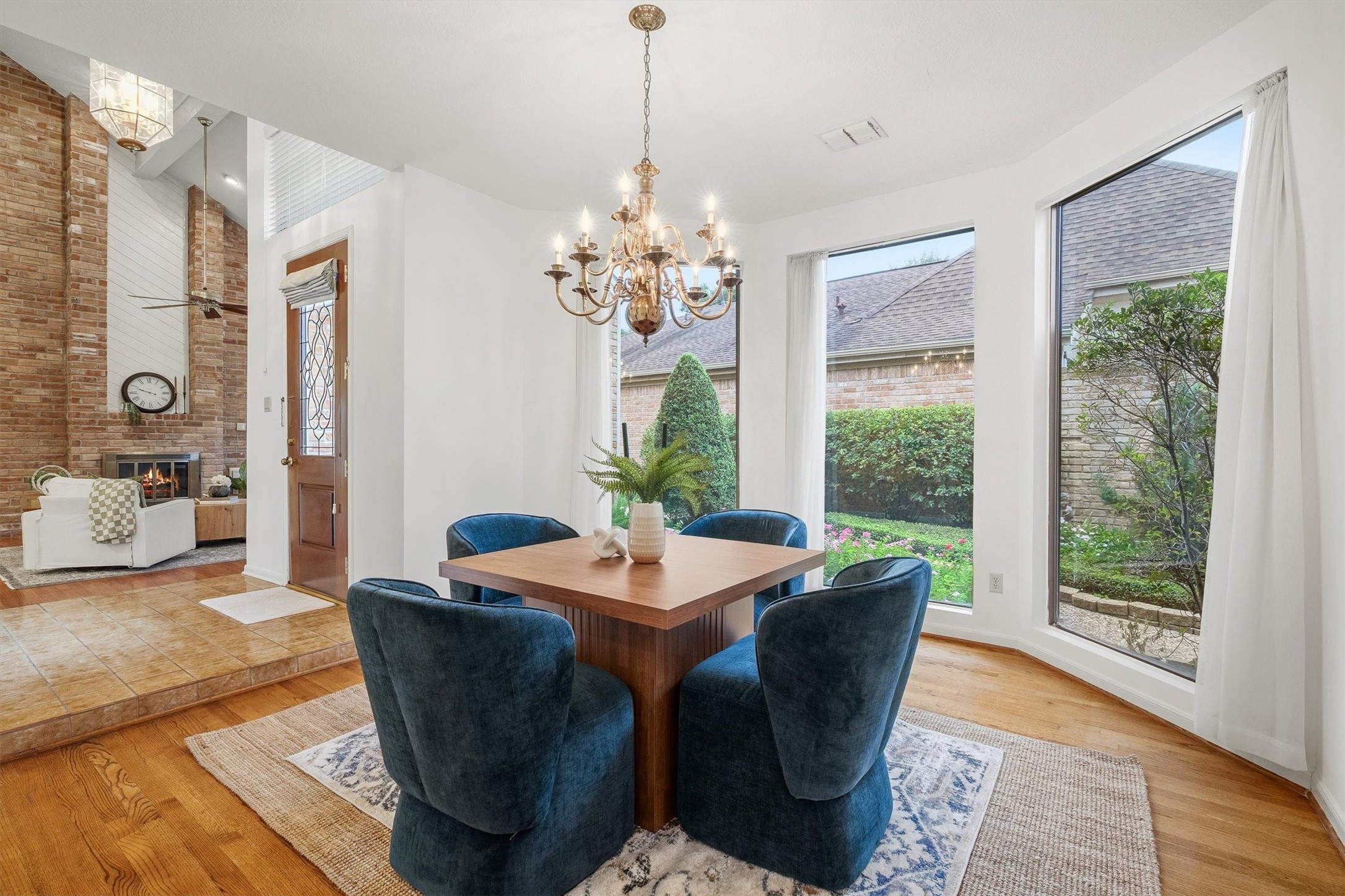 Great dining area with large windows providing garden views, complemented by a charming chandelier and plush seating. Adjacent is a cozy living space featuring a brick fireplace and high ceilings for an inviting atmosphere.