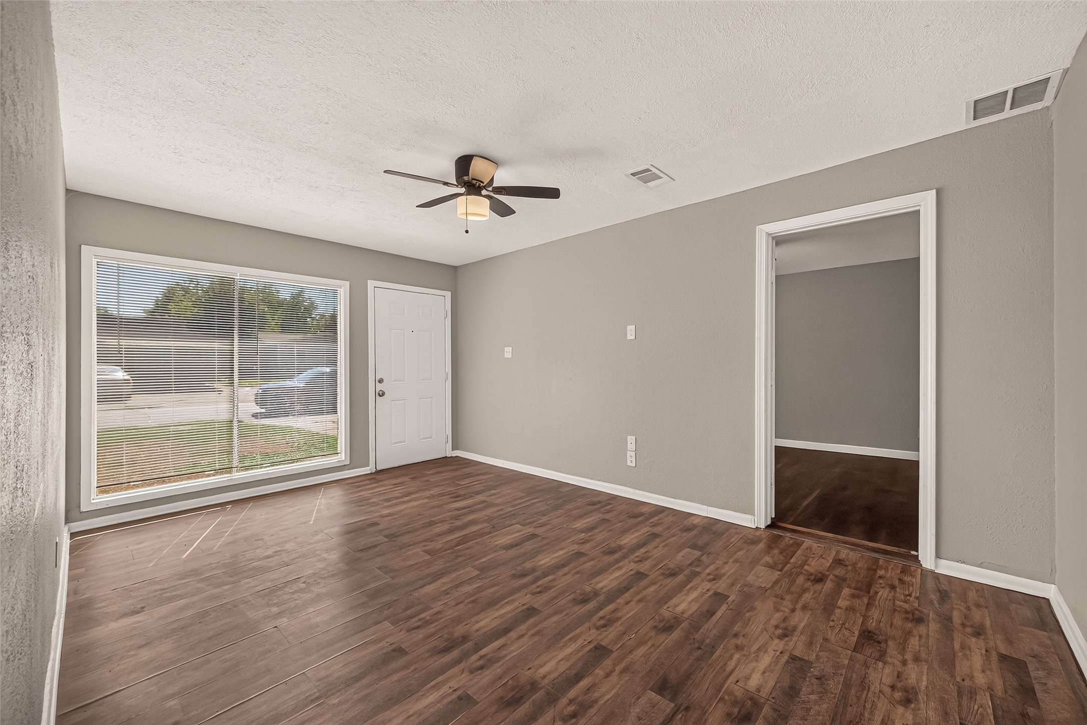 Living room, unfurnished, with large front window and ceiling fan.