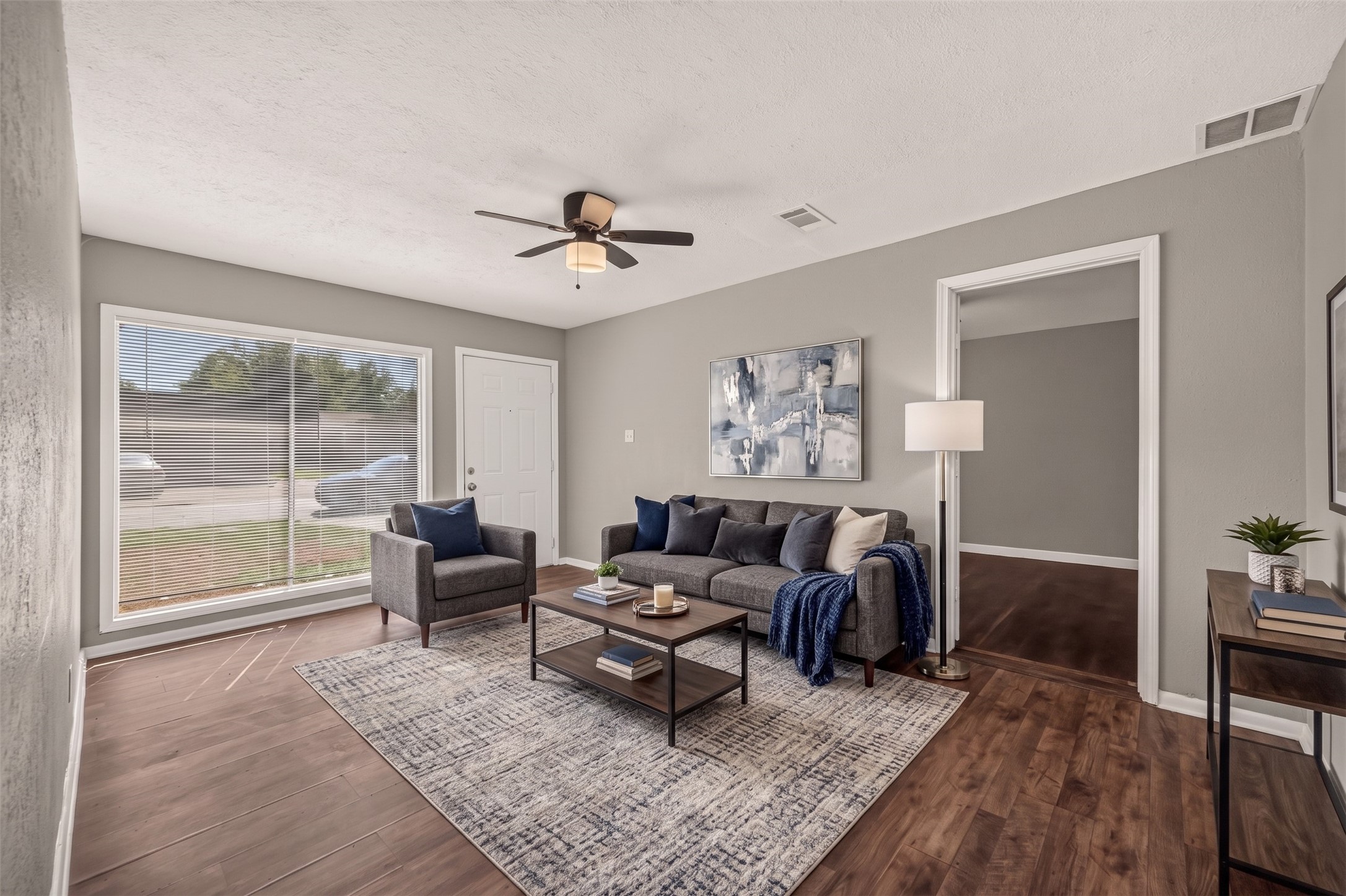 Living room with large front window and ceiling fan, open to the hallway. (Virtually staged)