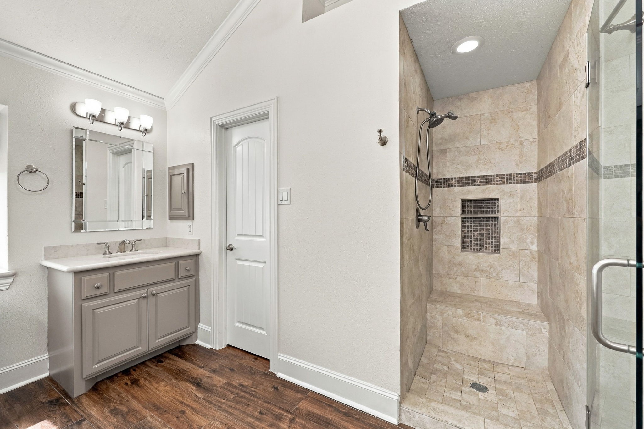 The large walk-in shower features a frameless glass door, a built-in bench, floor-to-ceiling tiled walls with an elegant accent strip, a brushed nickel showerhead and handheld wand, and convenient built-in niches for shampoo, soap, and other bath essentials.
