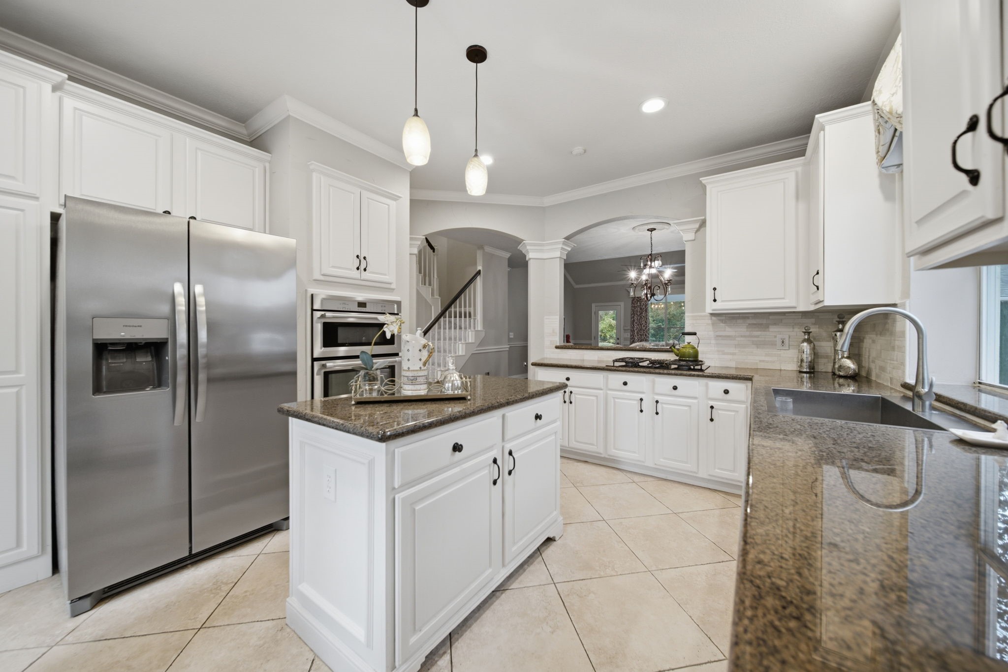 This elegant kitchen featuring white cabinetry, granite countertops, and stainless steel appliances including the refrigerator opens up to the living area. The space is complemented by a central island, stylish pendant lighting, and a spacious layout with ample natural light creating a welcoming setting for everyday living.
