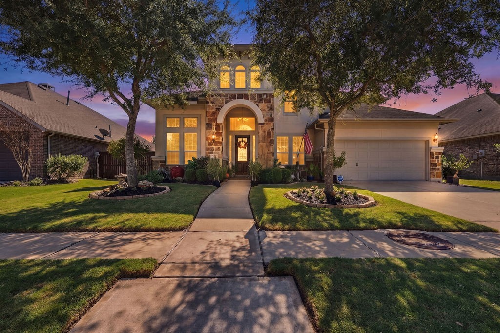 Stunning two-story home with elegant stone accents, illuminated windows, and a welcoming arched entryway, nestled on a beautifully landscaped lot with mature trees.