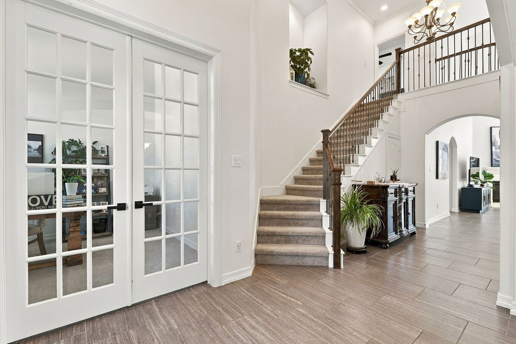 Elegant entryway featuring a grand staircase, stylish French doors leading to a cozy office, and modern tile flooring, all under a sophisticated chandelier.