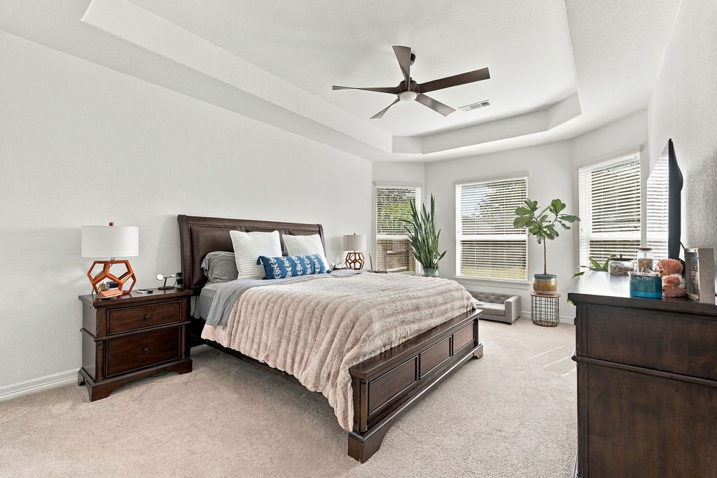 Spacious primary bedroom with a tray ceiling, large windows for natural light, and elegant decor featuring a cozy bed, stylish nightstands, and lush plants.