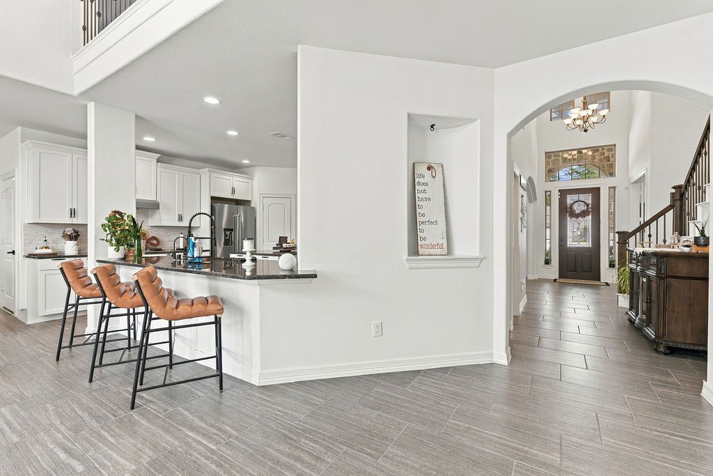 A modern open-concept kitchen and entryway featuring sleek white cabinetry, a stylish breakfast bar with room for multiple barstools, and elegant tile flooring leading to a welcoming front door.